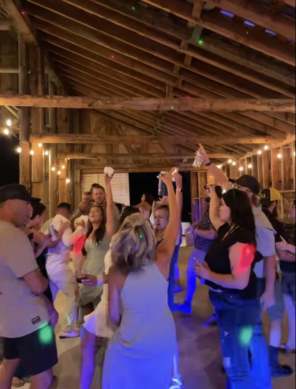 Group of people dancing and celebrating in a rustic wooden barn with string lights at night.