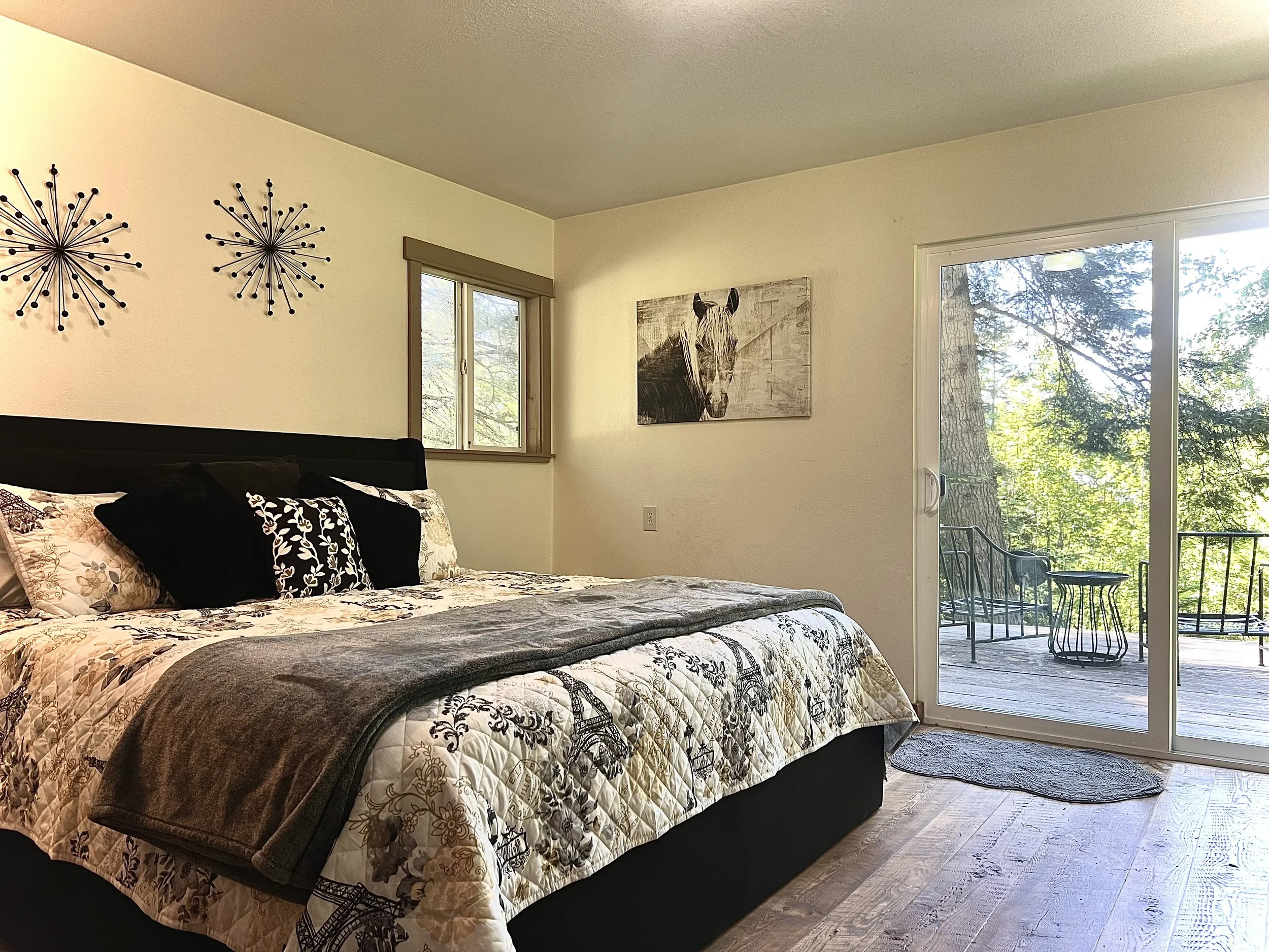 A bedroom with a bed covered in a floral and patterned quilt, black and floral pillows, two wall decorations resembling starbursts, a window, a painting of a horse on the wall, and sliding glass doors opening to an outdoor patio with chairs and trees outside.