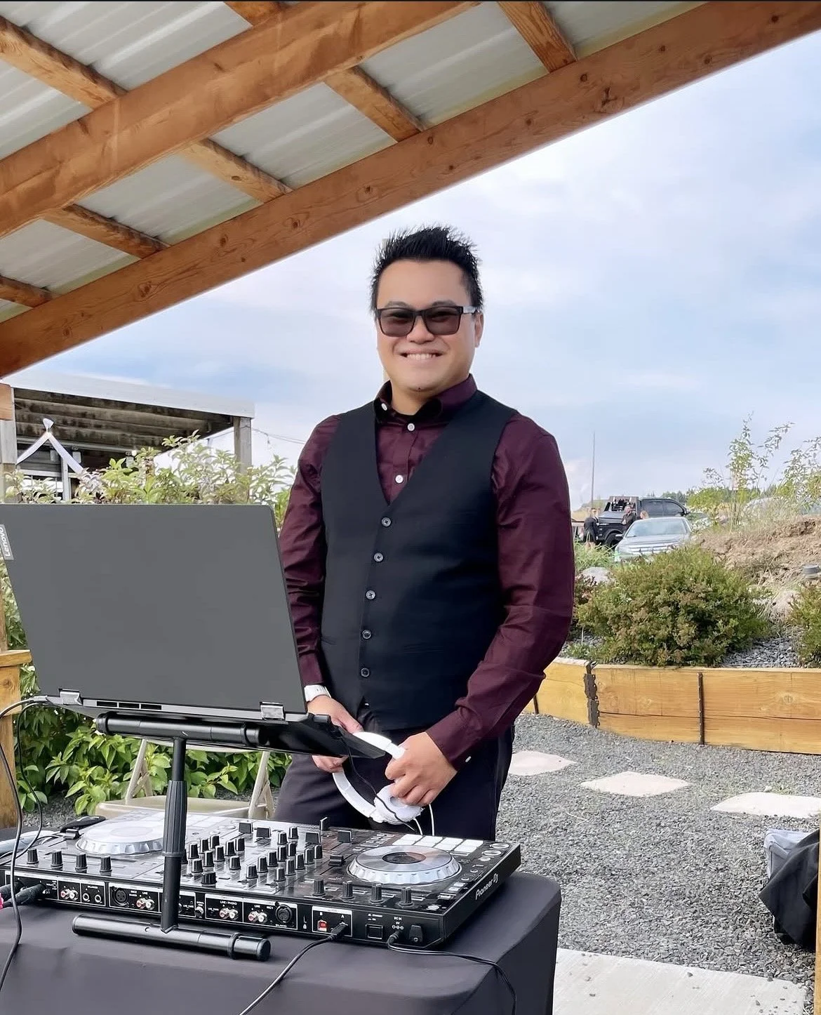 A man standing outdoors under a wooden shelter with a DJ controller and laptop, smiling at the camera, with parked cars and bushes in the background.