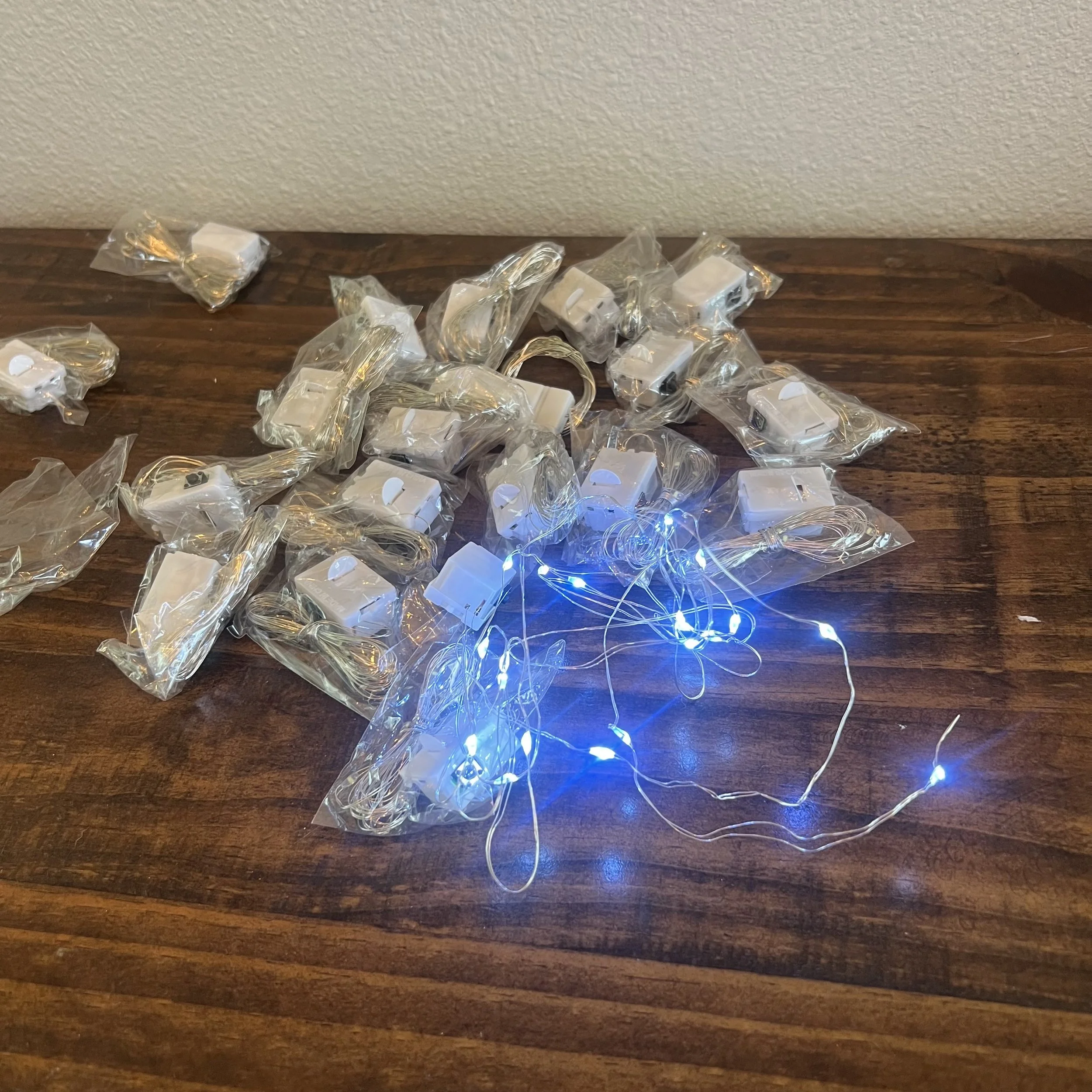 Multiple small white electronic devices and cables wrapped in clear plastic packaging are scattered on a dark wooden surface against a beige textured wall.