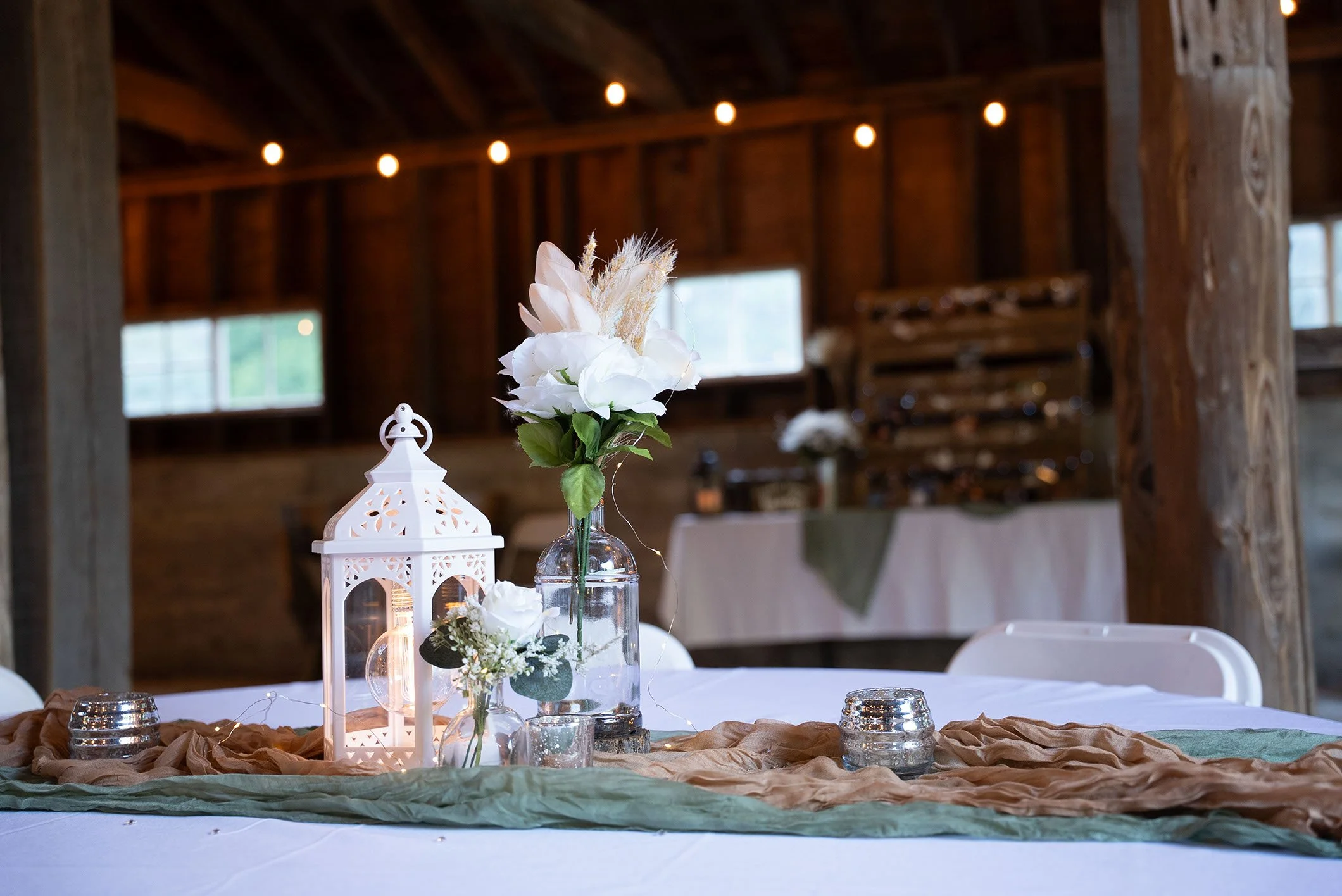 Wedding reception table centerpiece with white flowers, a lantern, and candles in a rustic wooden venue.