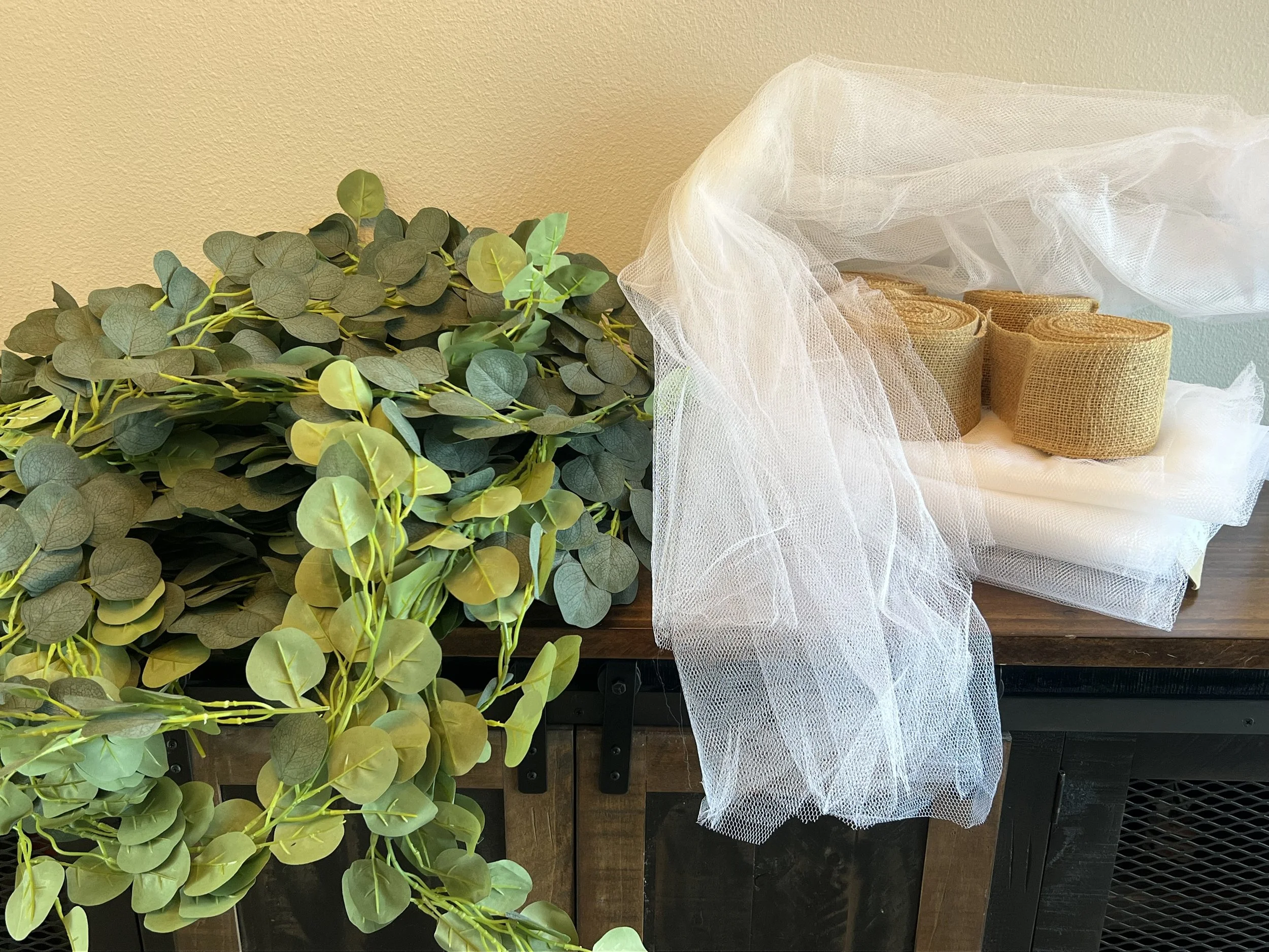 Artificial green plant on table with burlap-wrapped scrolls and white tulle fabric.