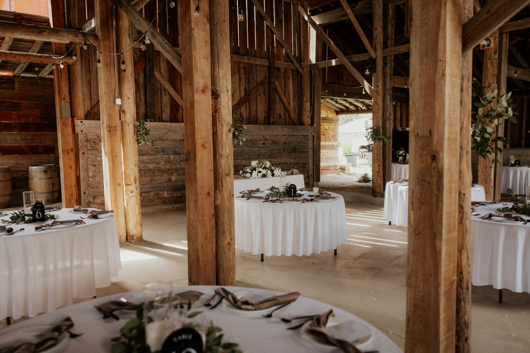 Wooden indoor wedding reception area with round tables draped in white tablecloths, decorated with greenery and place settings. String lights hang from the ceiling, and wooden beams and walls create a rustic atmosphere.