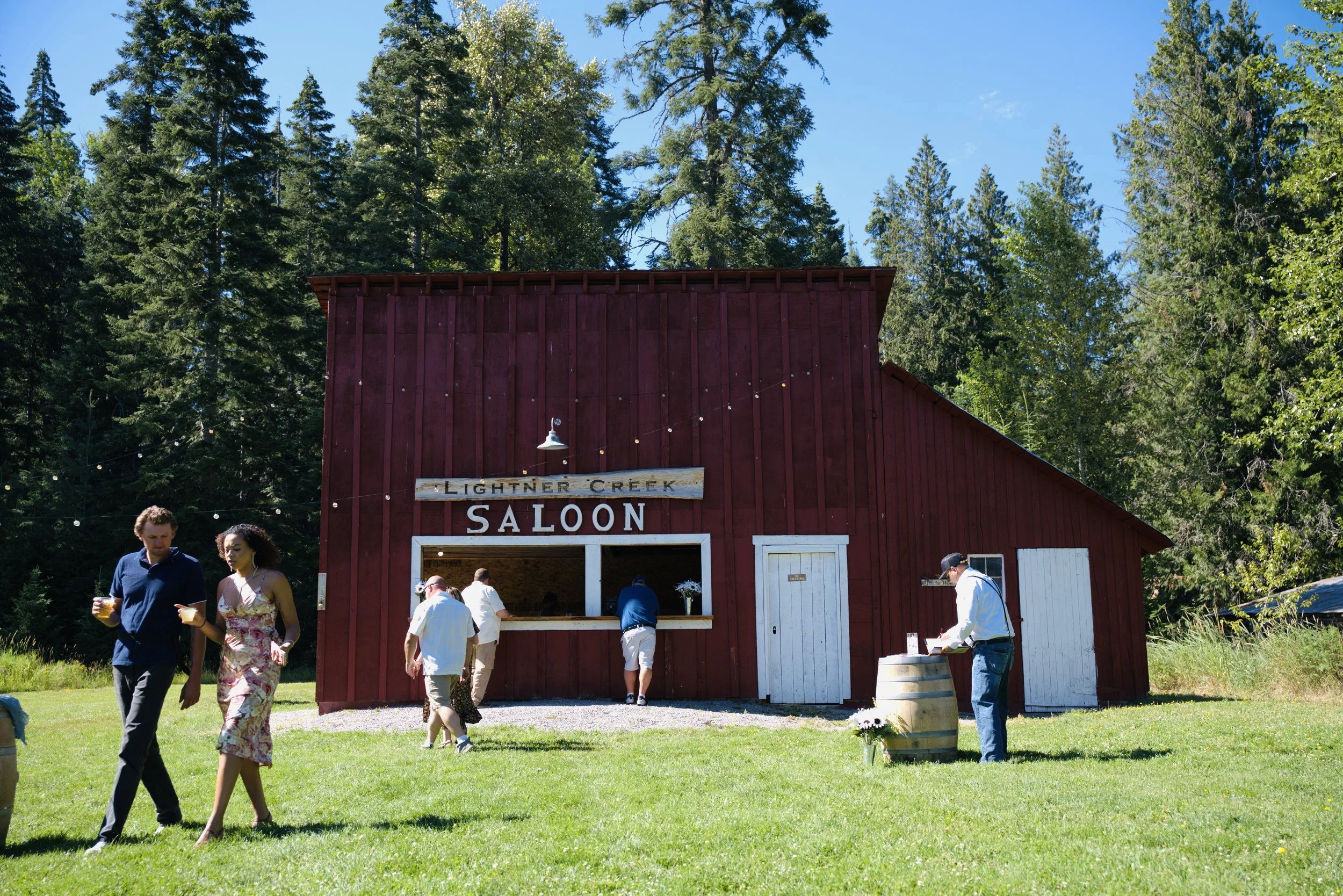 People gather outside a red barn-style building labeled 'Lighter Creek Saloon' on a sunny day. Trees and a clear blue sky are in the background.
