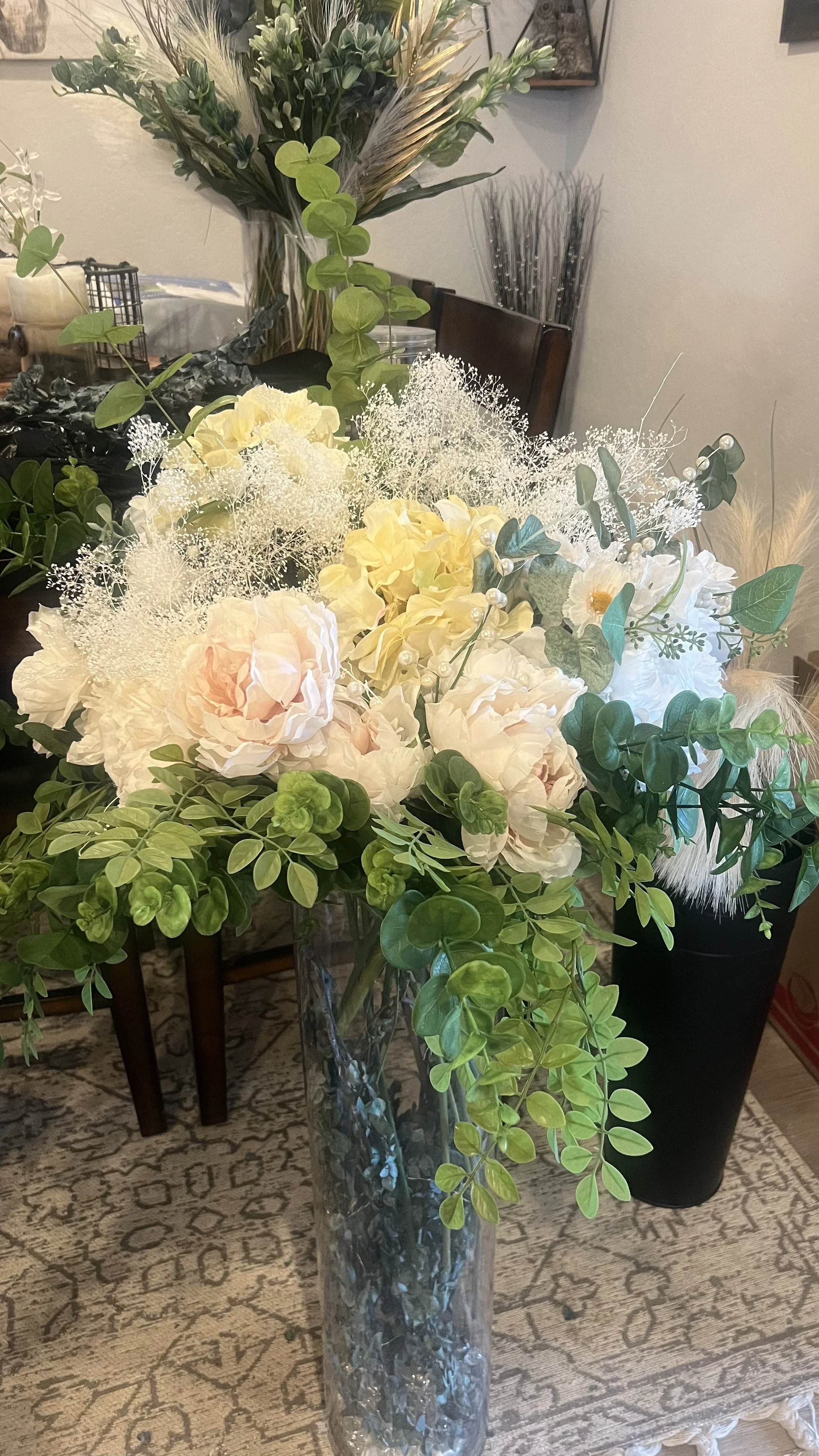 A tall glass vase with a floral arrangement of white and pale pink flowers, green leaves, eucalyptus, baby's breath, and other greenery in a living room or dining area.