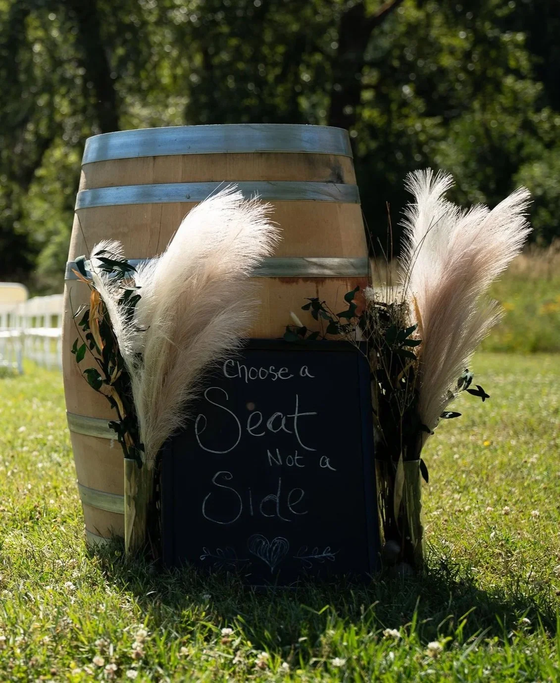 A wooden barrel with metal bands outdoors on grass, decorated with white pampas grass and greenery, and a small chalkboard sign that says 'Choose a seat, not a side' with a heart drawn underneath.