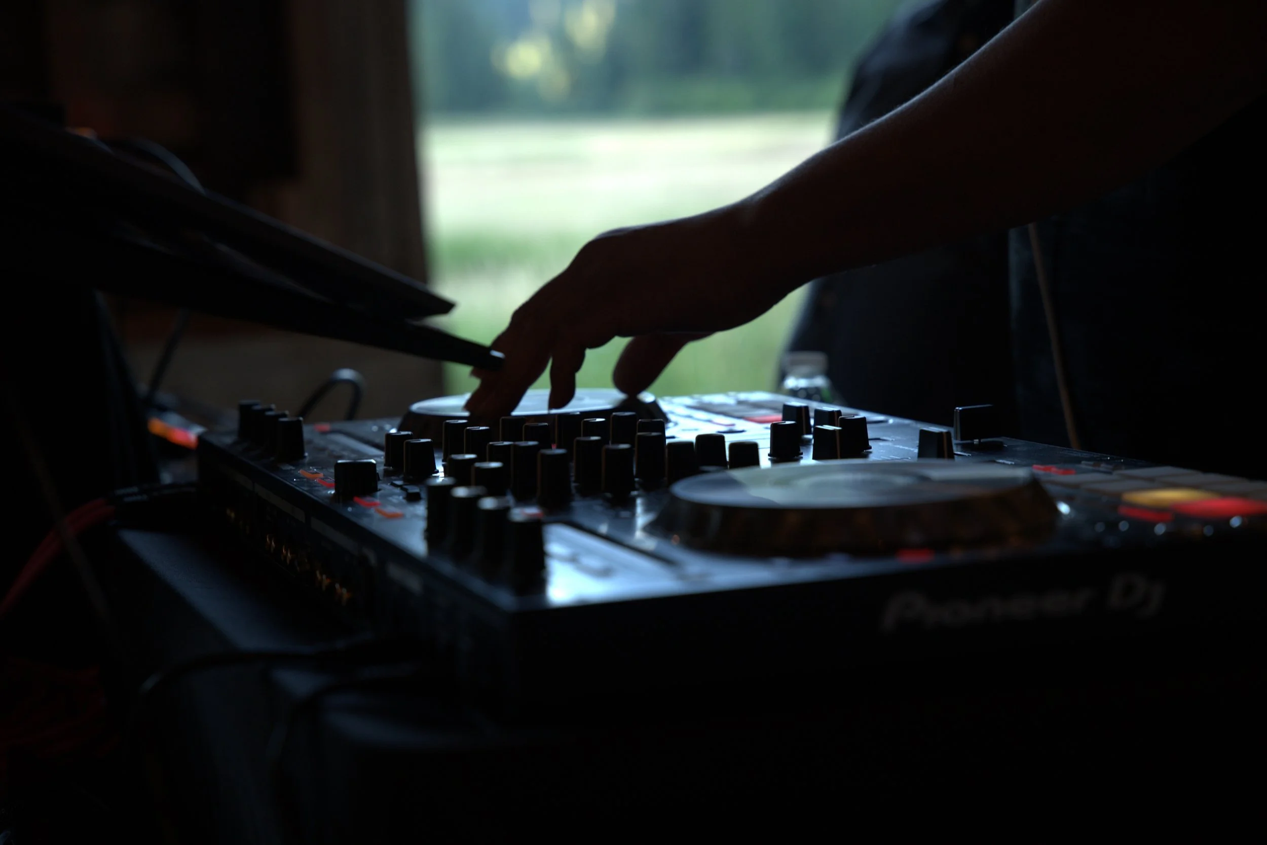 A person’s hand operating a DJ mixer or turntable with a blurred outdoor background visible through a window.