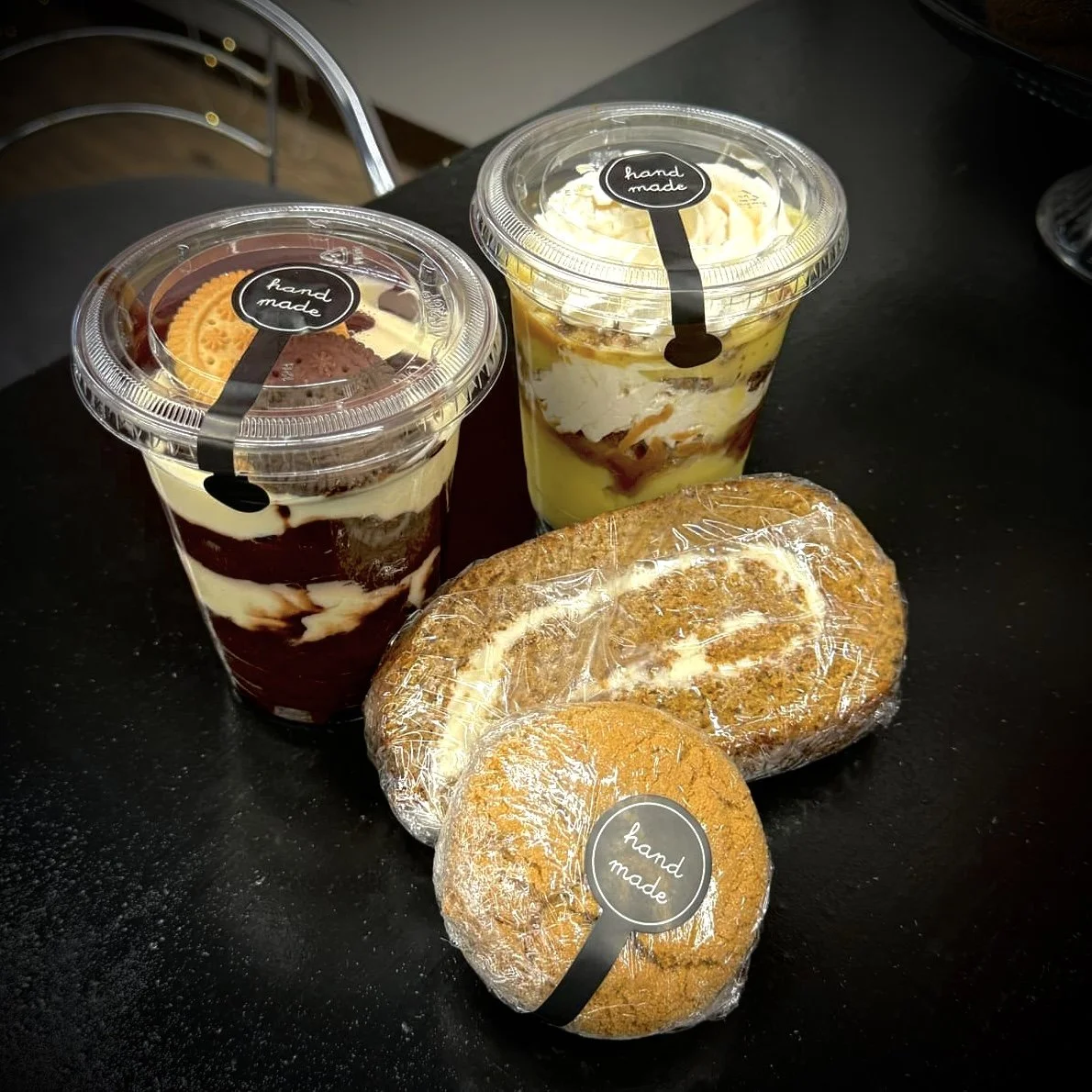 Two cups of layered desserts with 'hand made' labels, a packaged pastry bread, and a round muffin with a 'hand made' label on a black surface.