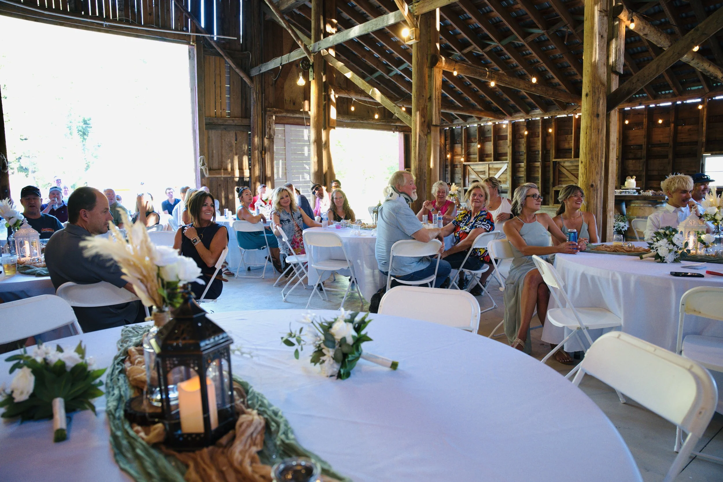 Indoor rustic barn wedding reception with guests sitting at decorated tables, string lights, and natural sunlight coming through open doors.