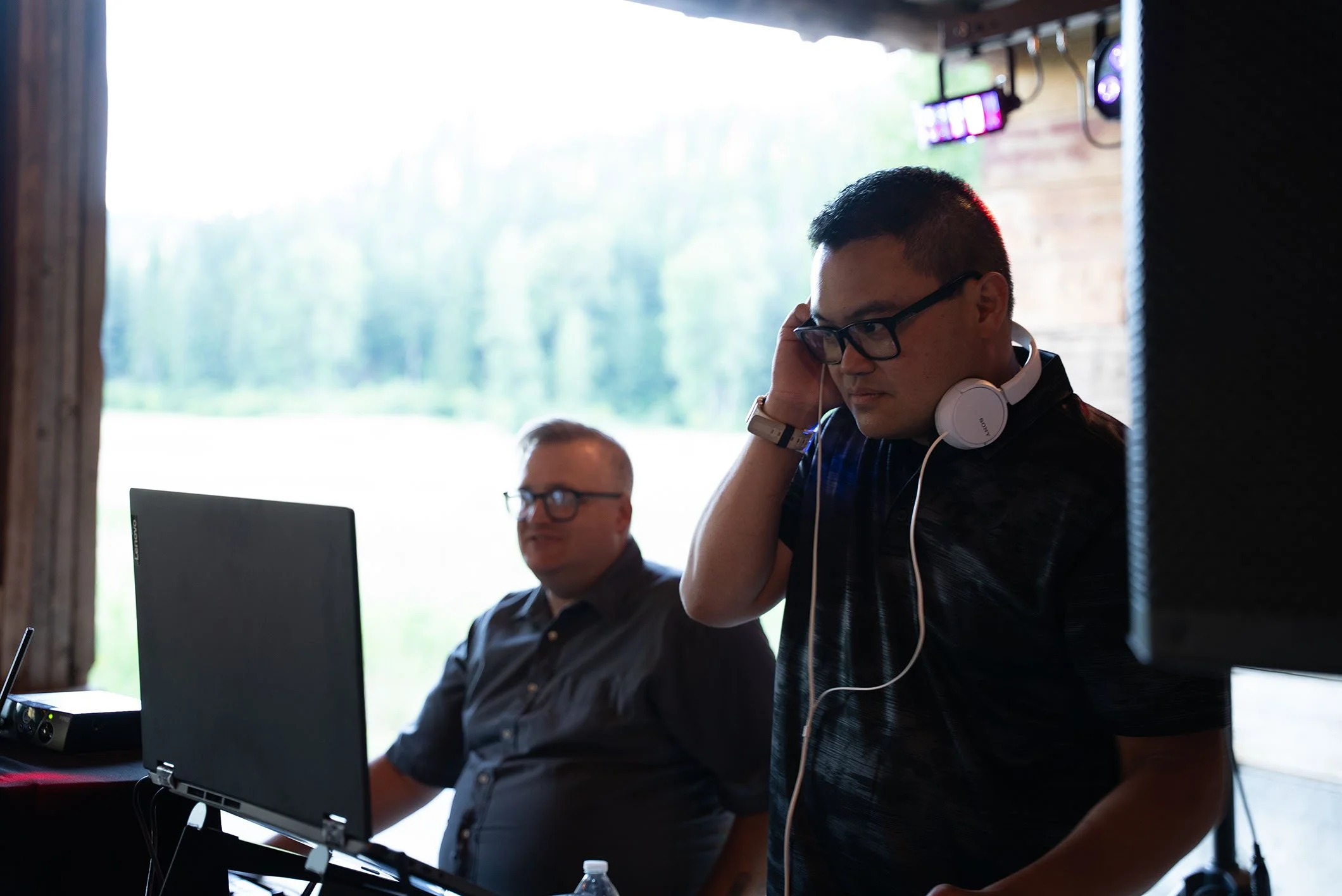 A man with glasses and a black shirt adjusting his white Sony headphones while standing near a seated man with glasses working on a laptop in a room with large windows and trees outside.