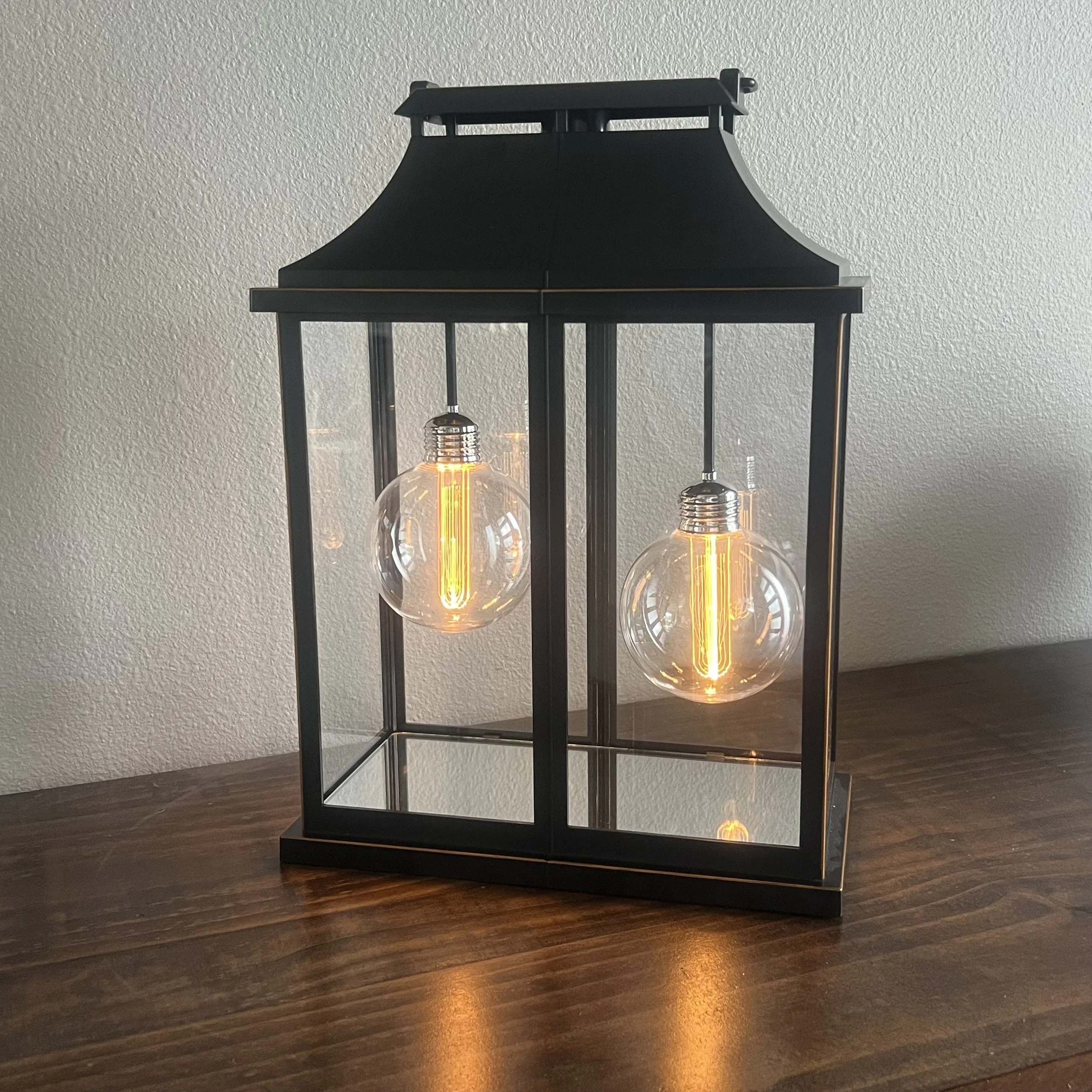 A black metal lantern with two exposed vintage-style light bulbs inside, placed on a wooden surface against a white textured wall.