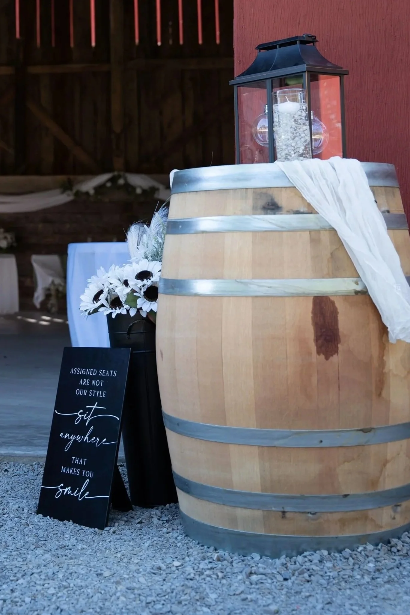 A wooden barrel with metal bands and a lantern on top, decorated with a white cloth. There is a sign that reads, "Assigned seats are not our style, sit anywhere that makes you smile." A bouquet of white flowers is visible behind the sign, and the setting appears to be an indoor or outdoor event space with rustic decor.