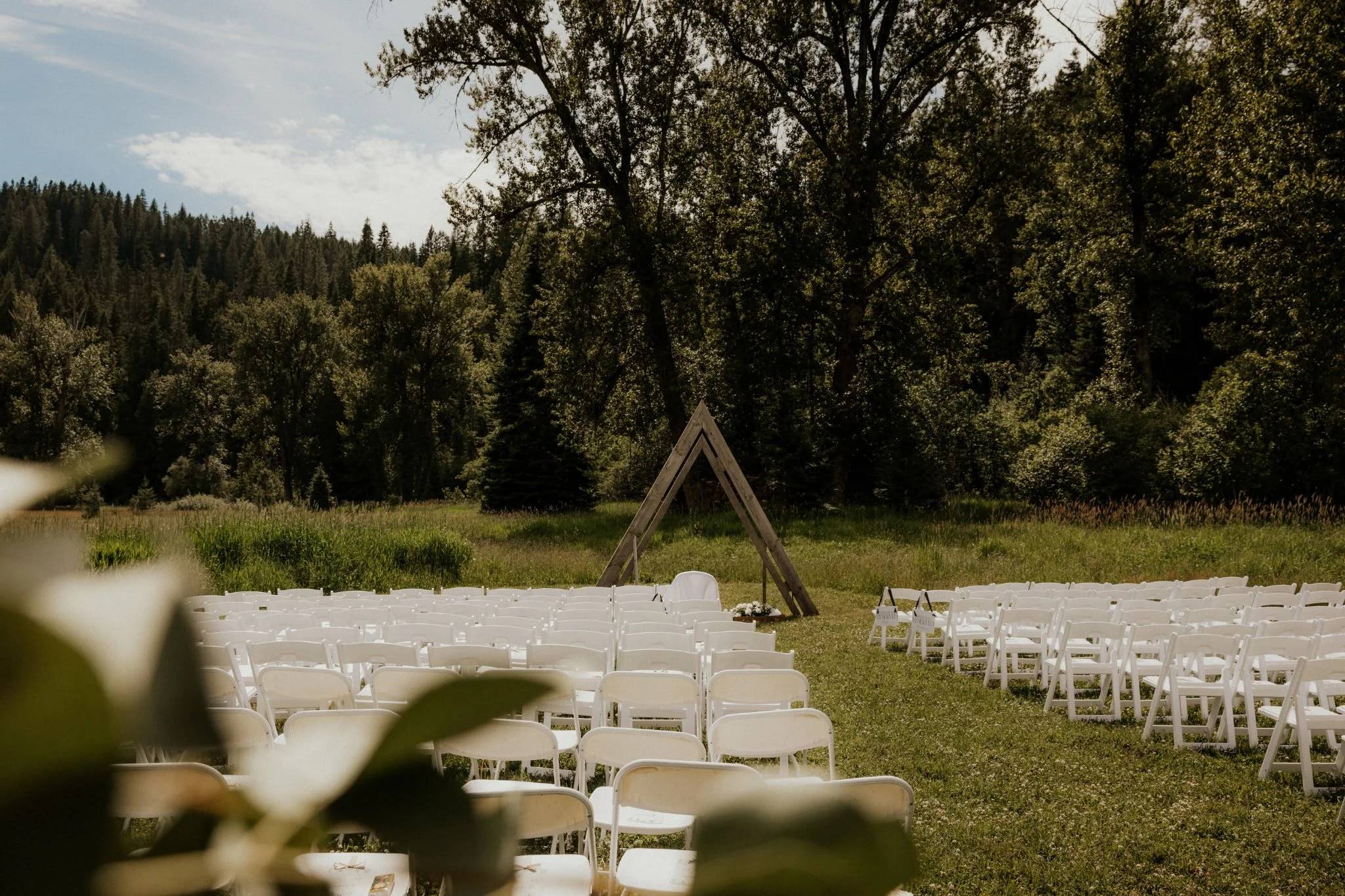 Outdoor wedding ceremony setup with white chairs arranged in rows on grass, a wooden triangular arch in front, and a scenic forest backdrop under a partly cloudy sky.