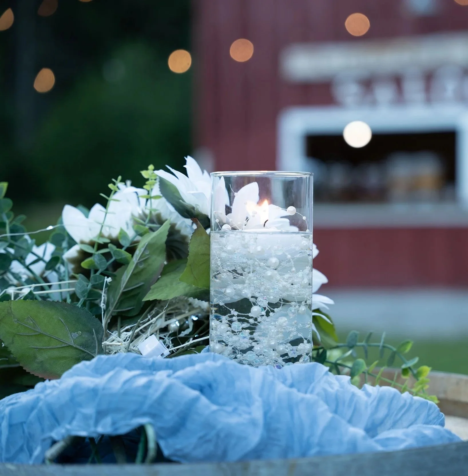 Close-up of a lit white candle inside a glass holder, surrounded by white and green artificial flowers, with blurred warm lights in the background.