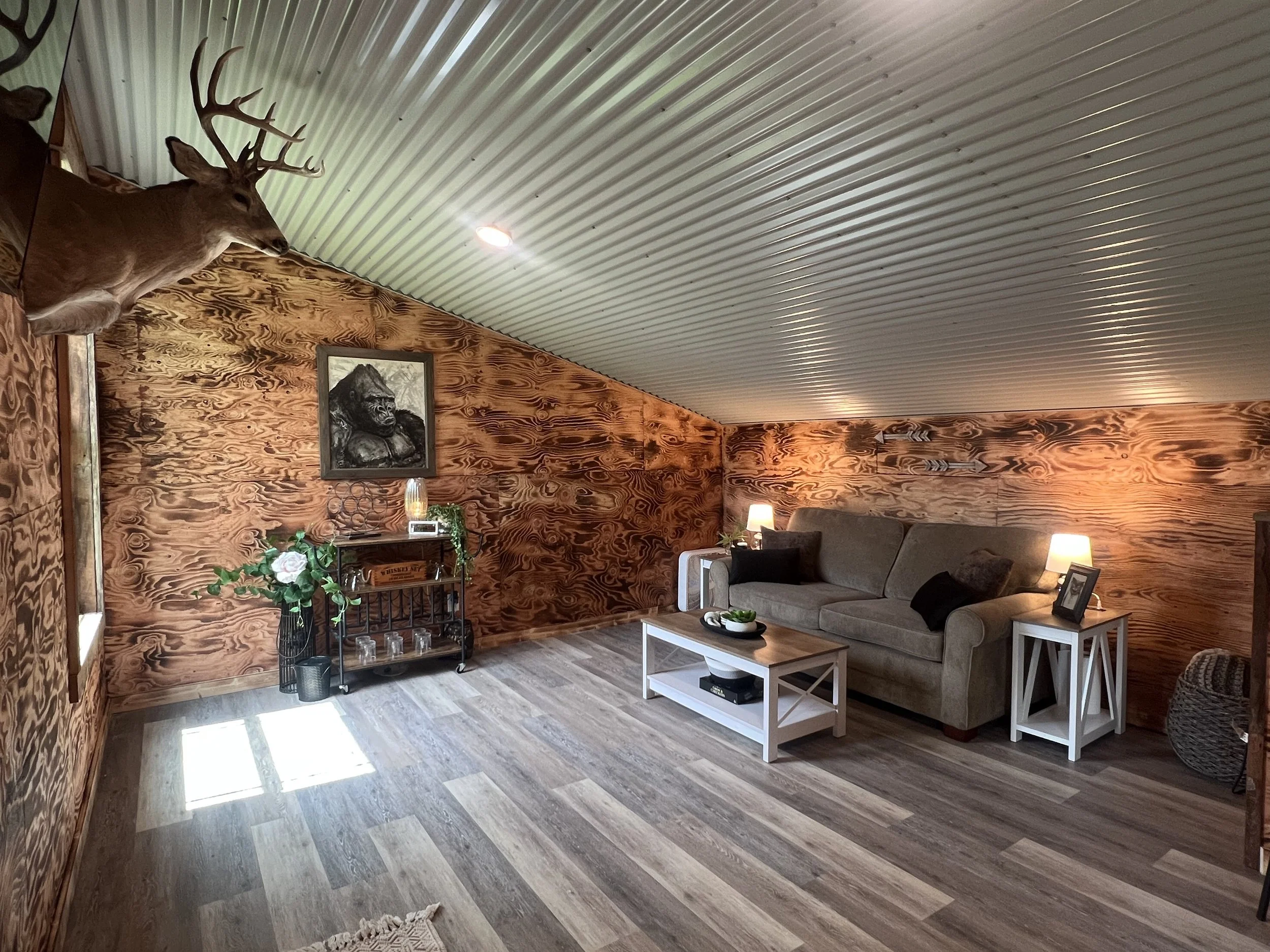 Living room featuring wood-paneled walls, a mounted deer head, a framed gorilla artwork, a gray sofa with black and white pillows, white side tables, a black metal cart with decorative items, lamps, and a plant on a stand.