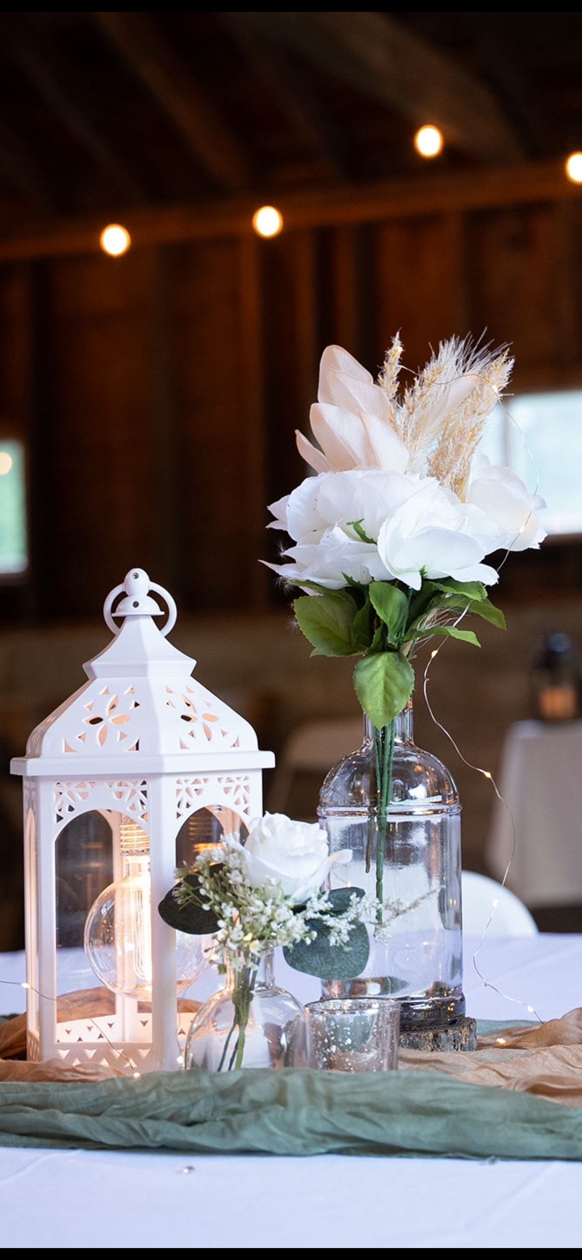 A table centerpiece with white flowers in a glass vase, a white decorative lantern, and string lights in a rustic indoor setting.