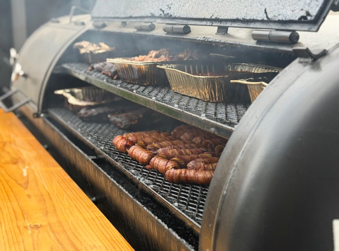Meat and vegetables grilling inside a large smoker or grill with a glass window, placed on a wooden table.