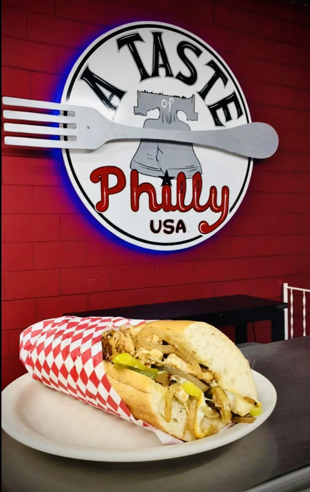 A hot dog with grilled onions and pickles in a red and white checkered paper wrapper on a white plate, set against a red brick wall. Behind, a neon sign reads "Taste of Philly USA" with a fork and a circle.