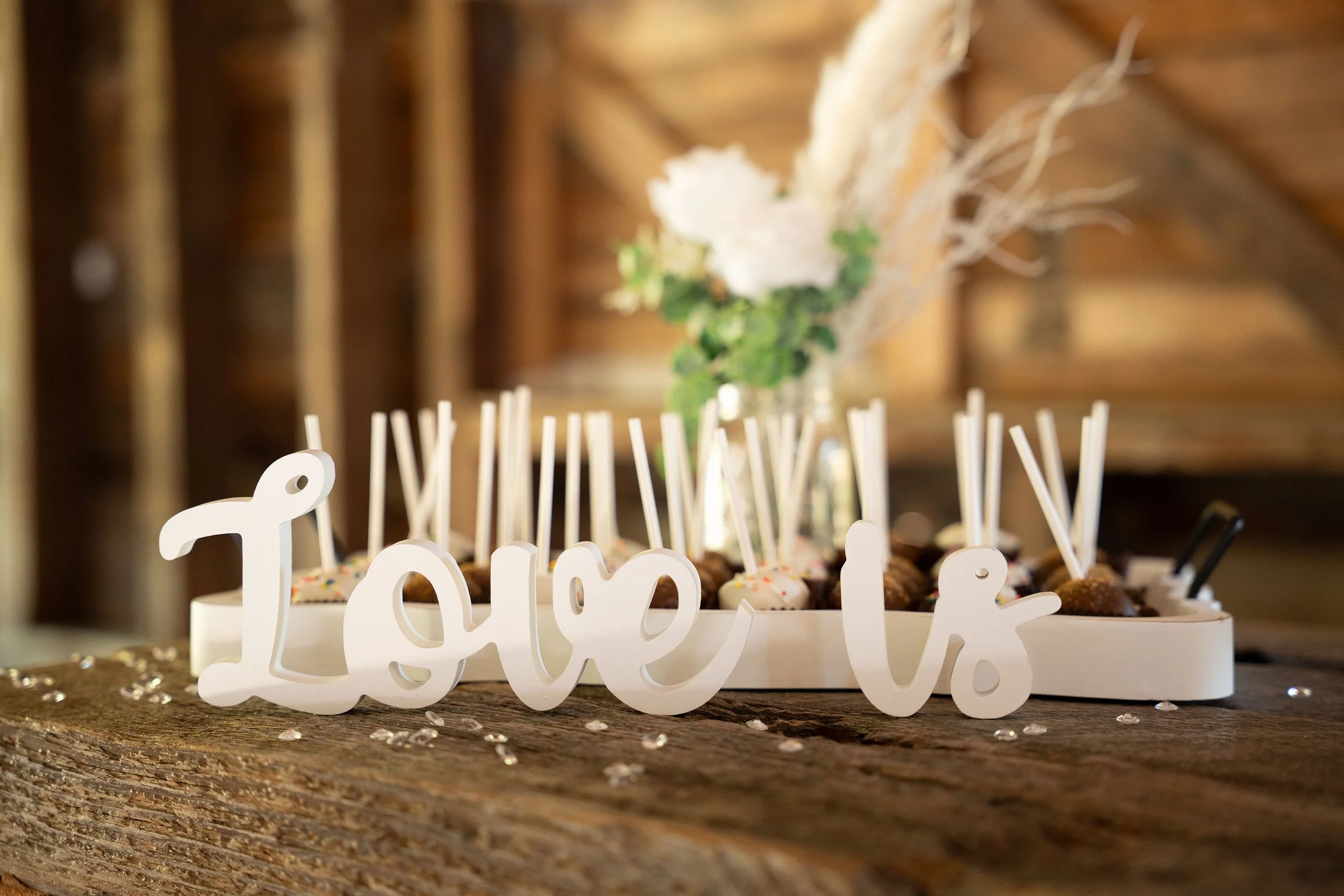 A birthday cake with a white "happy" cake topper, decorated with colorful sprinkles and small cake pops with white sticks. In the background, there are blurred flowers in a glass container and a wooden wall.