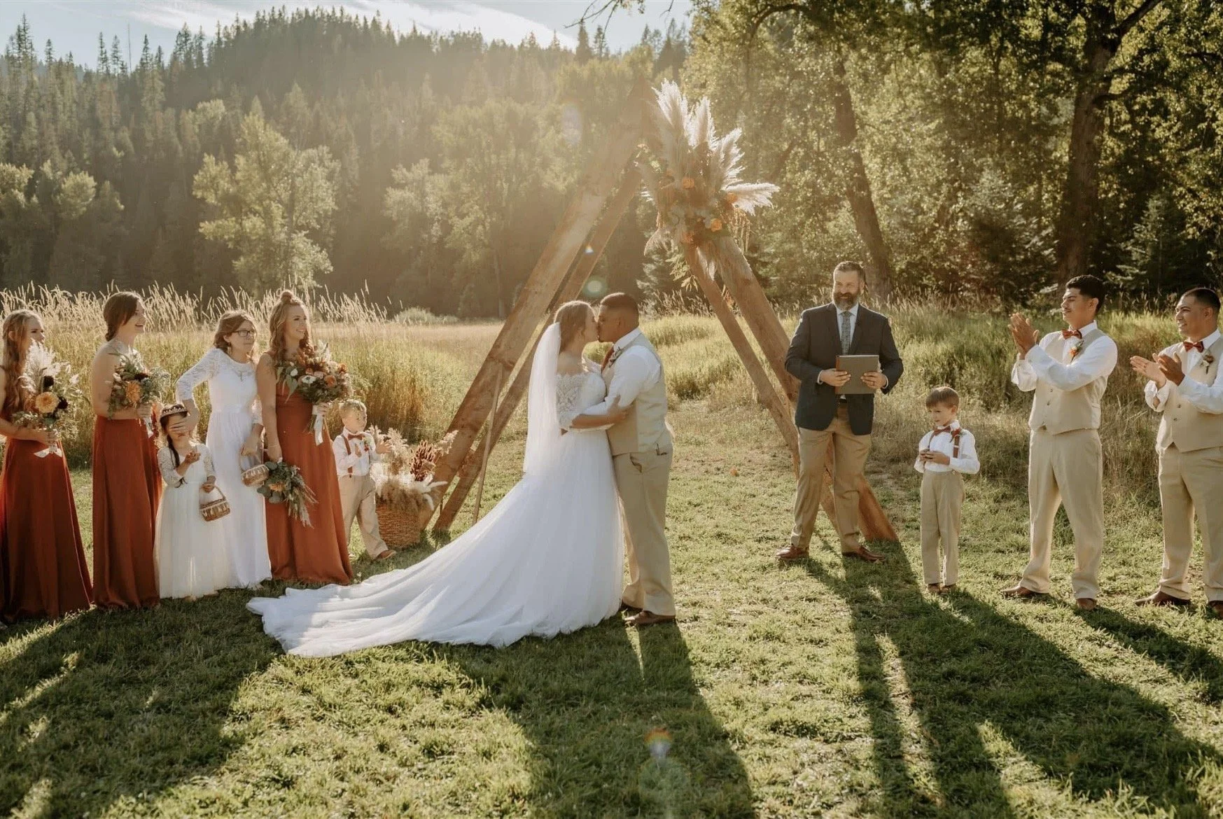 A couple kisses at their outdoor wedding ceremony in a grassy field with a wooden arch decorated with flowers. Bridesmaids and groomsmen, including children, stand on either side, with the bridesmaids holding bouquets and the groomsmen clapping, all set against a background of trees and mountains in the sunlight.