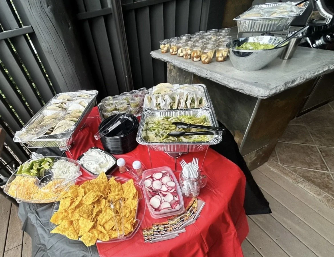 Buffet table with nacho chips, radishes, guacamole, sour cream, and various condiments, with salads and catering trays on a red and black tablecloth, set against a black fence backdrop.