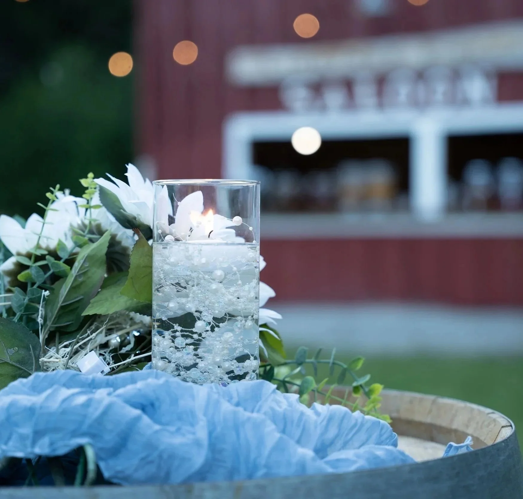 Close-up of a drinking glass with a candle and decorative white flowers, with blurred outdoor lights and building in the background.