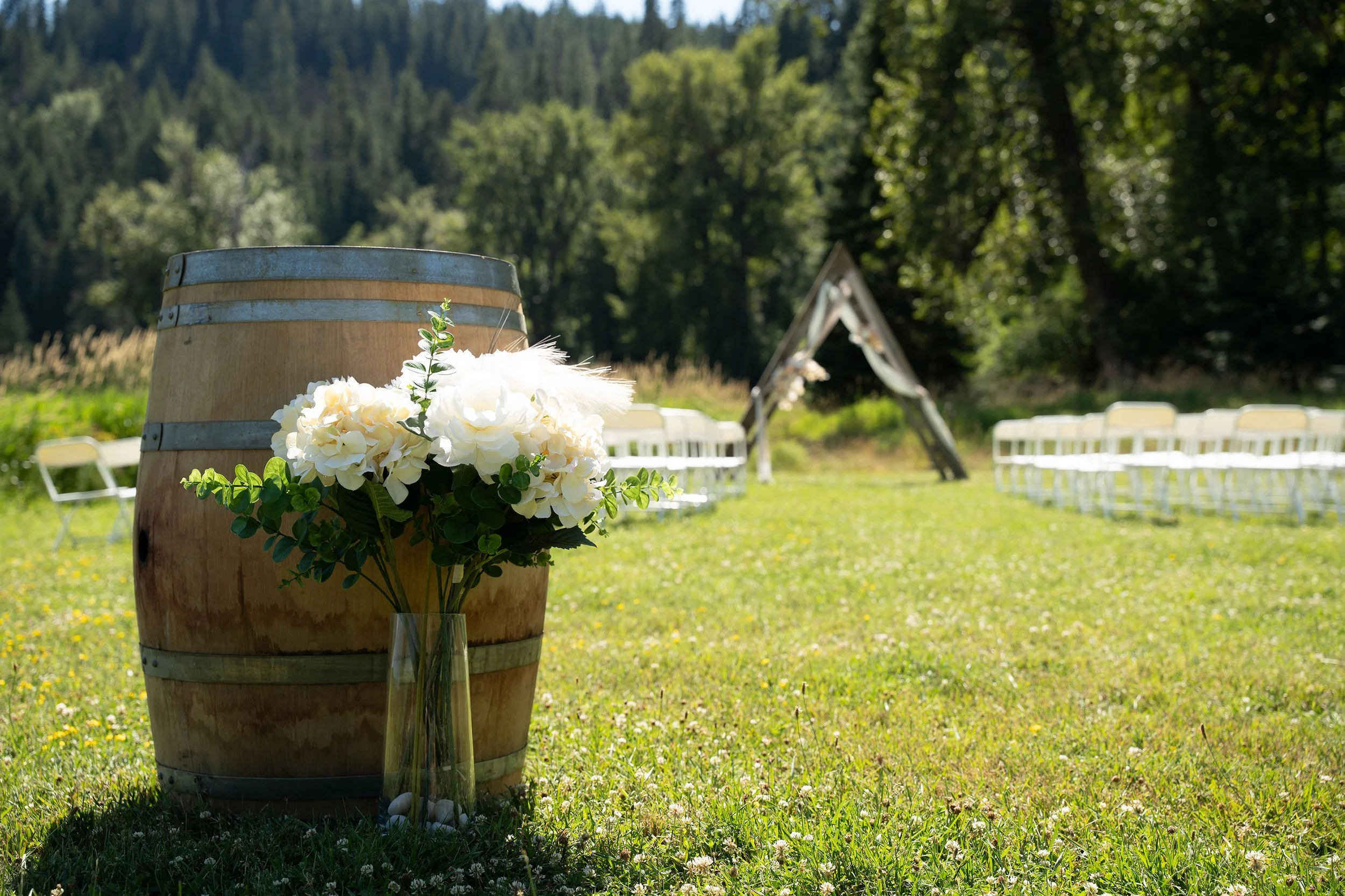 A wedding setup outdoors featuring a large wooden barrel with a bouquet of white flowers and greenery, with white chairs arranged in rows in the background, under trees and surrounded by greenery and mountains.