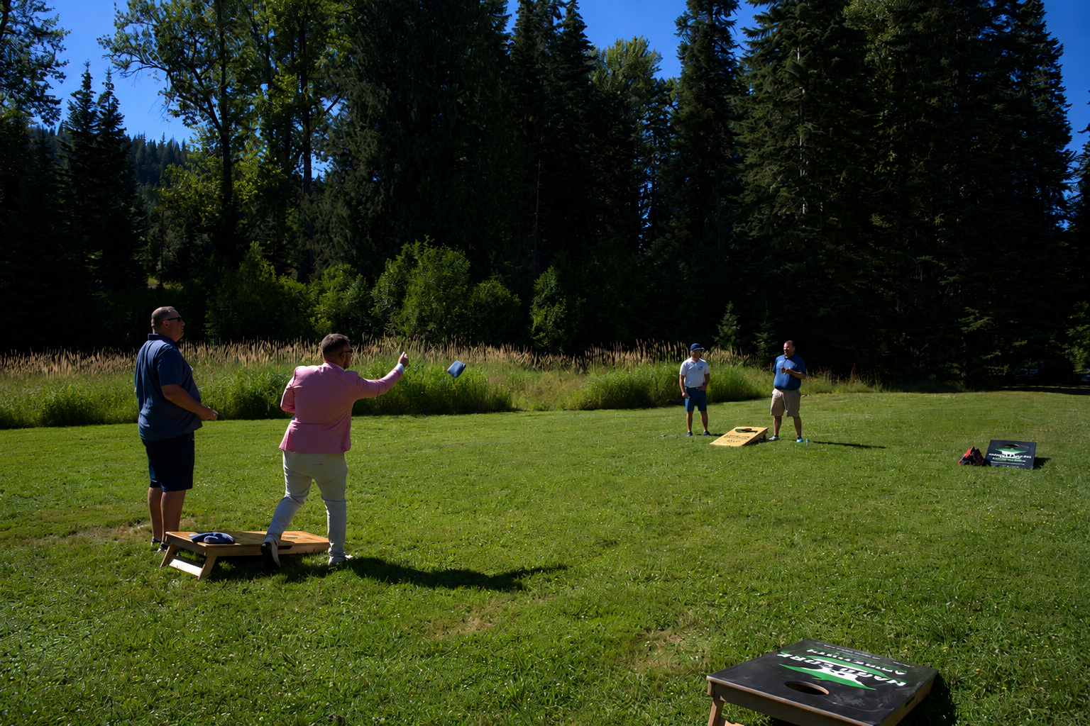 Four people playing cornhole outdoors on a grassy field surrounded by tall trees on a sunny day.