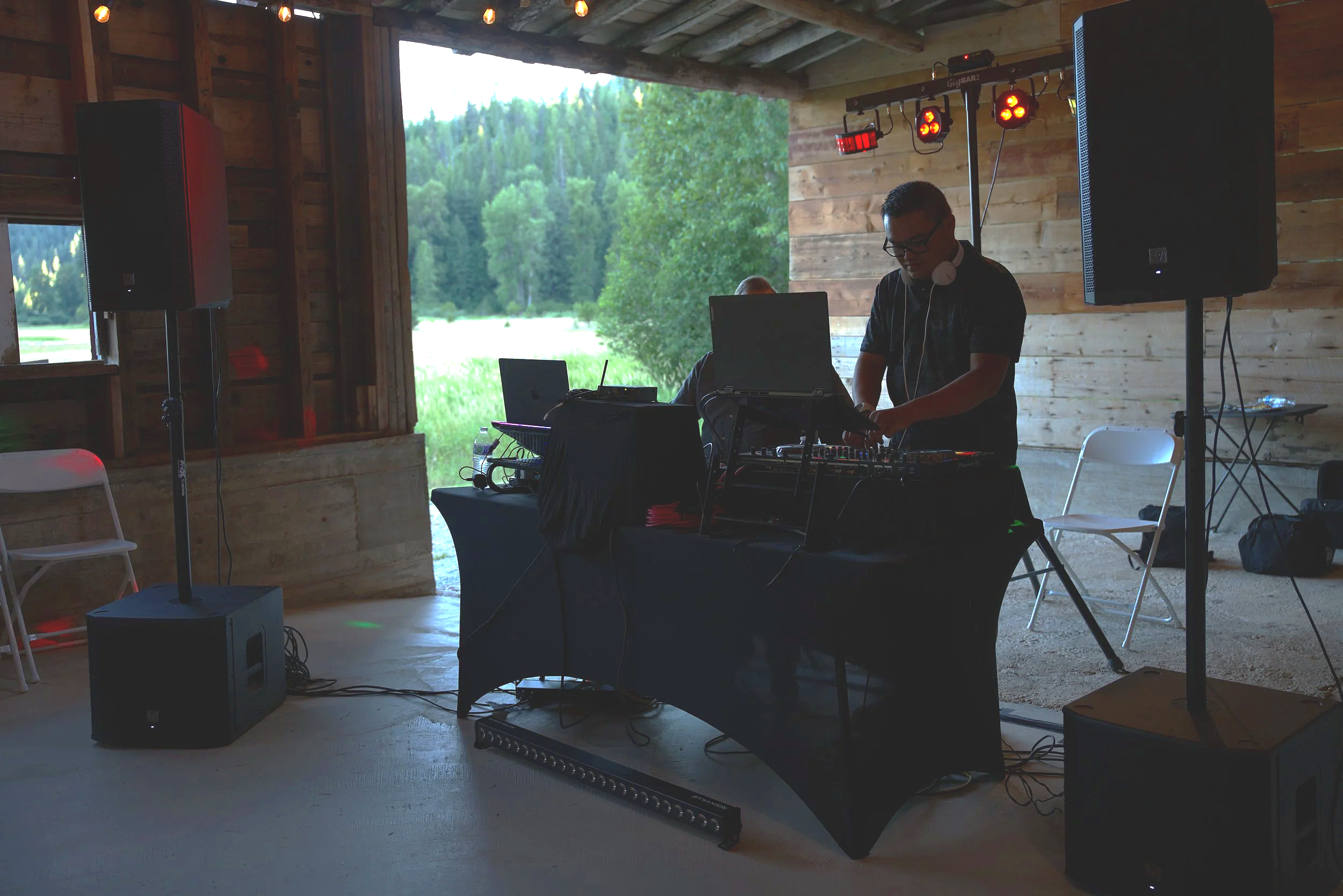 Man DJing at an indoor event with large speakers, audio equipment, and colored stage lights, with a view of trees and greenery through large open windows.