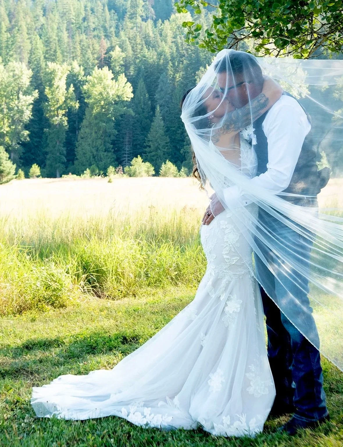 A bride and groom sharing a kiss outdoors with green trees and grass in the background. The bride is wearing a white wedding dress with floral embroidery and a long train, and a veil. The groom is in a suit and glasses.