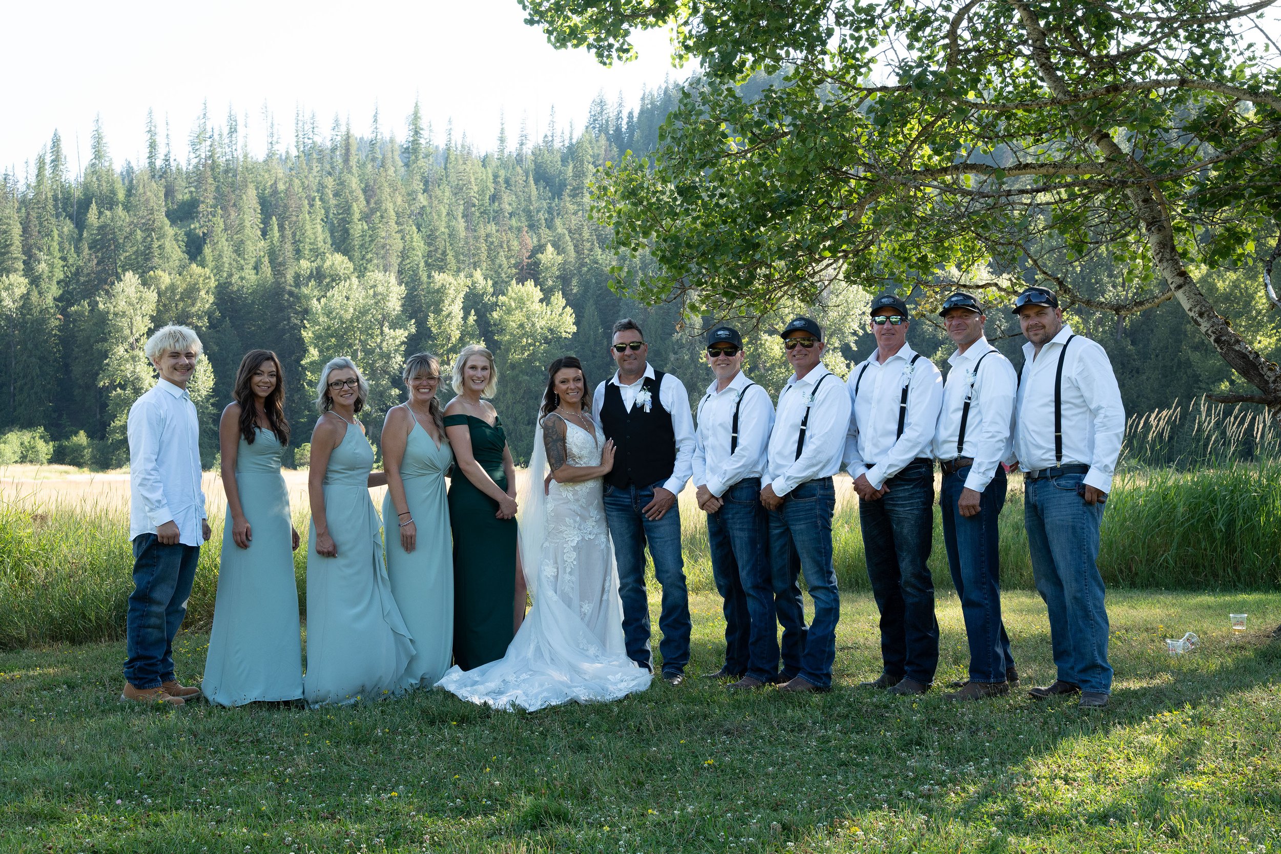 Group of people, including a bride and groom, standing outdoors in a grassy field with trees and a forest in the background, smiling and dressed in wedding attire and casual clothing.