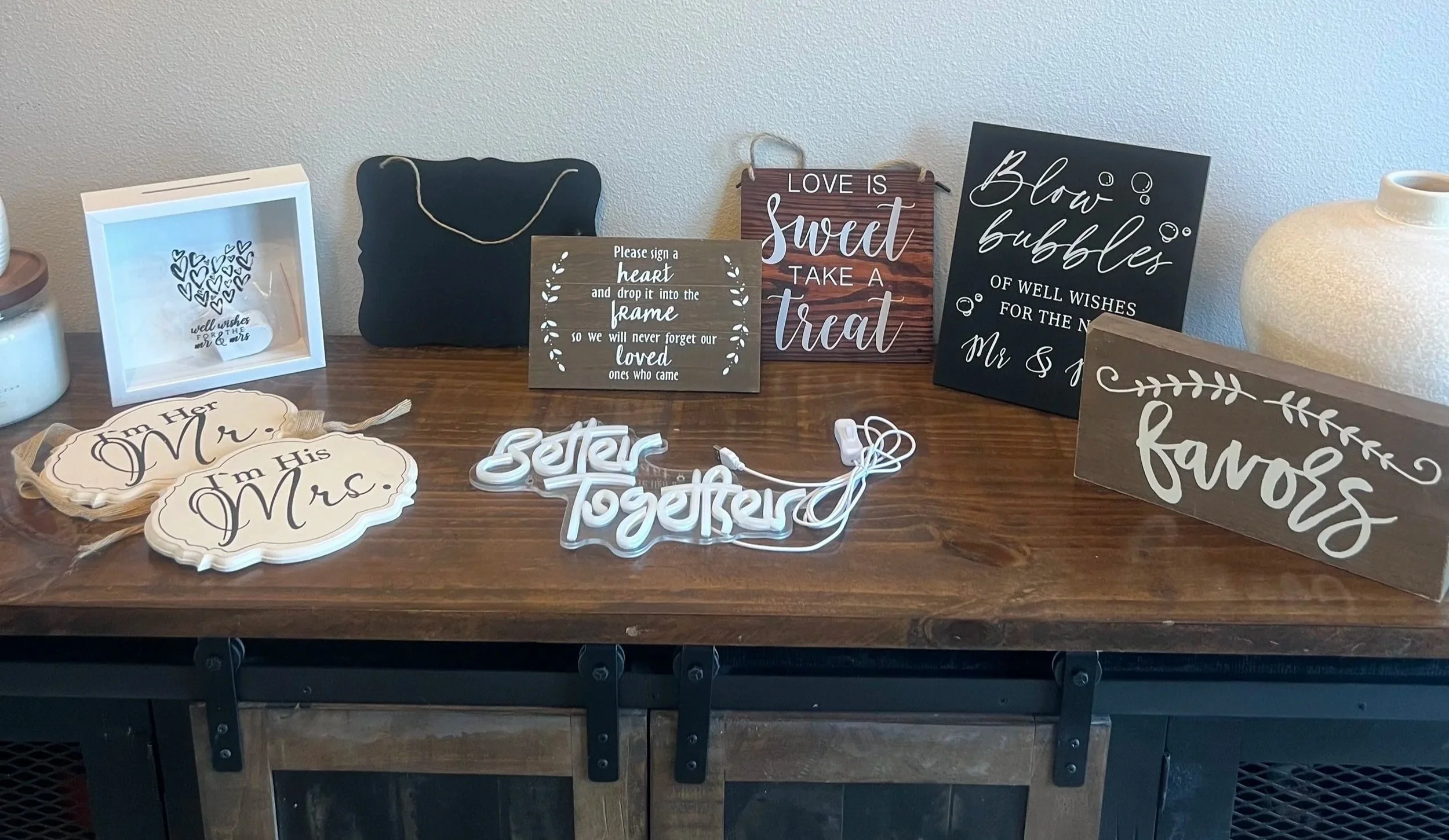 Gathering of wedding or event decorations on a wooden table, including signs with messages about love and marriage, a white shadow box, a black bag, heart-shaped signs that say 'I'm Her Mrs.' and 'I'm His Mr.', a decorative word sign that says 'Together', and a ceramic vase.