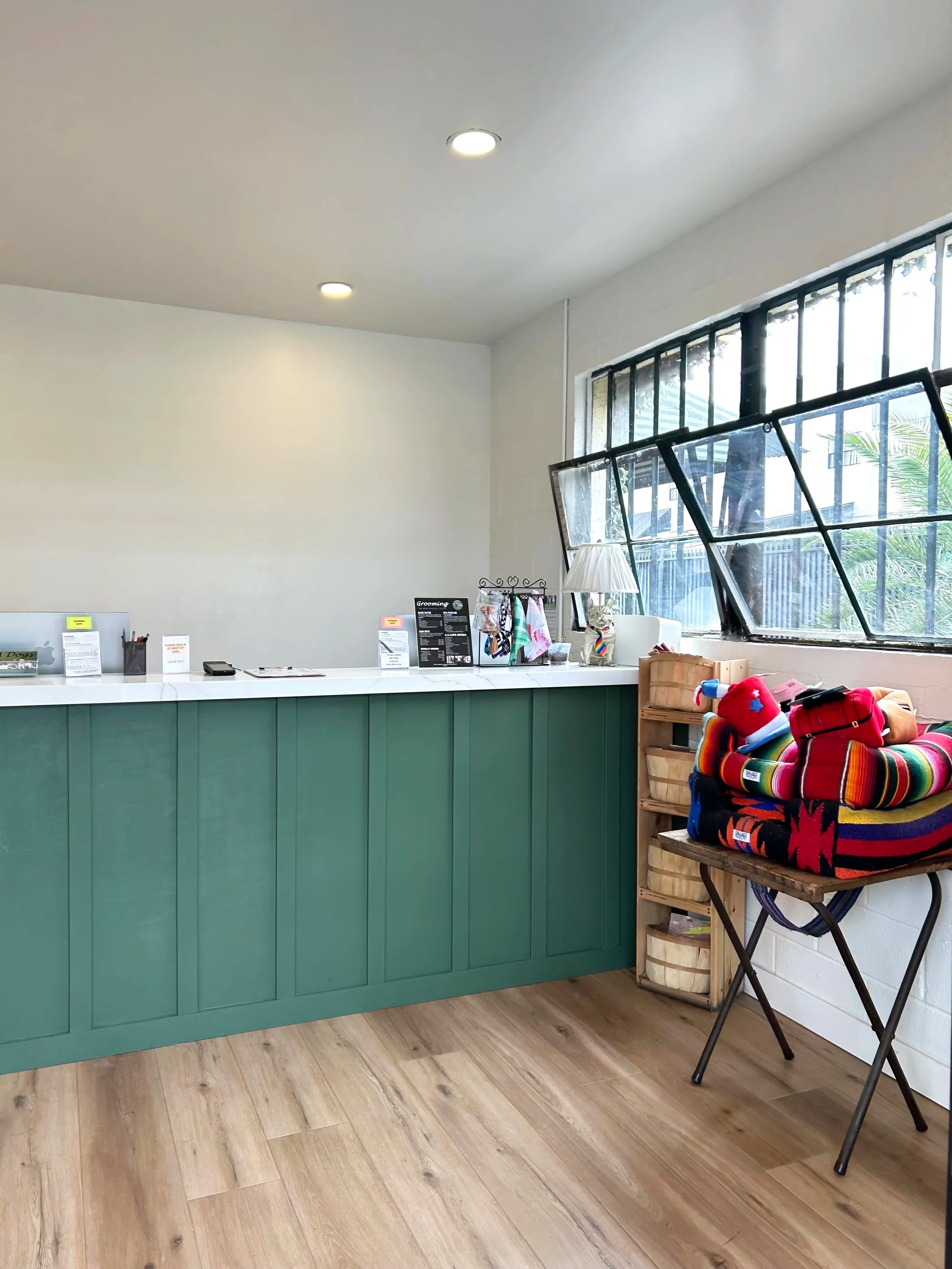 Interior of a room with an L-shaped white counter with green lower cabinets. There are two windows with black bars, one of which is open. Items on the counter include a small lamp, notices, and grooming products. To the right, there’s a wooden tray with colorful blankets or clothing, possibly pet supplies, on a small table. The room has wooden flooring and a white wall with ceiling lights.