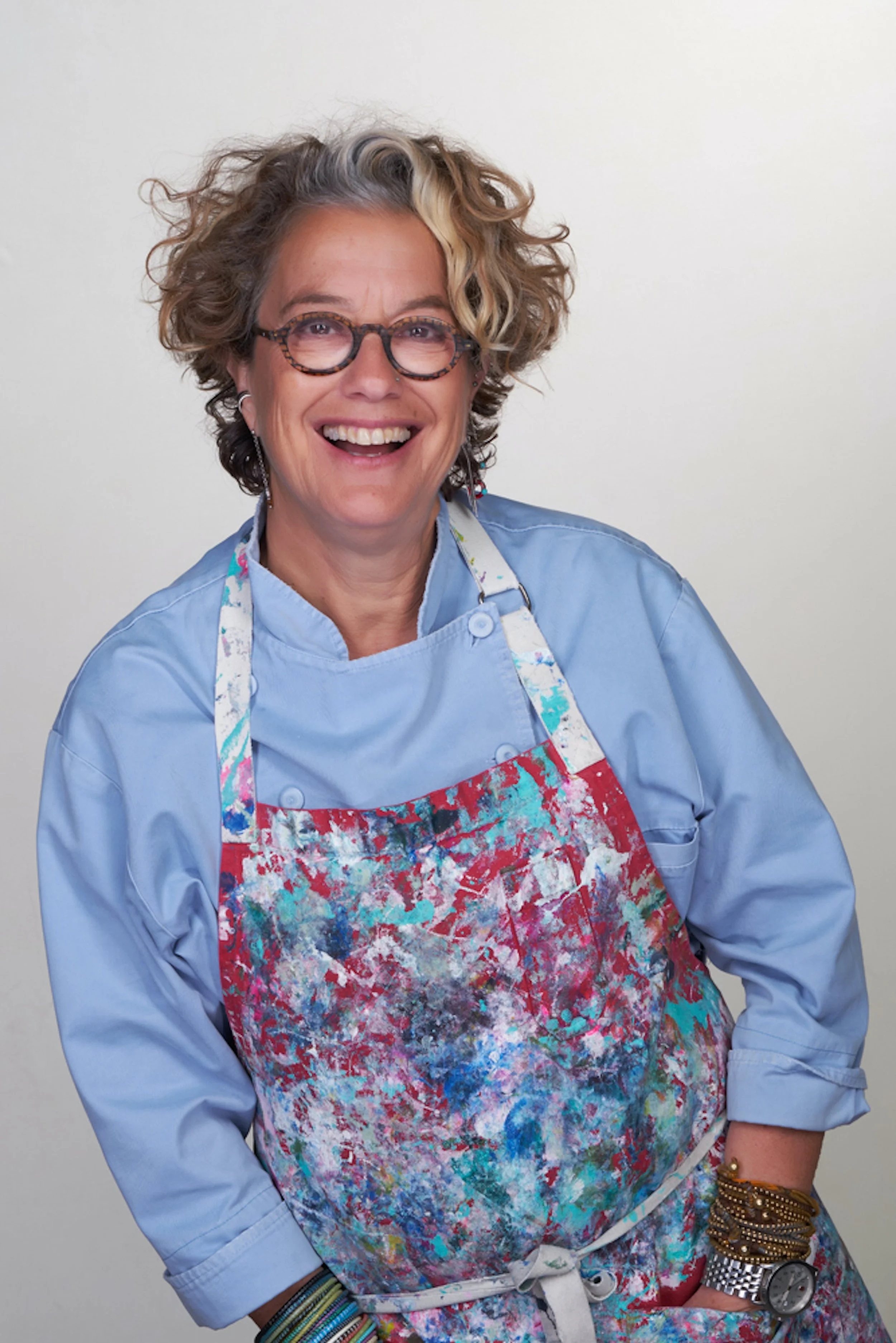 Smiling woman with curly hair, glasses, and multiple bracelets, wearing a light blue shirt and a colorful apron, standing against a plain background.
