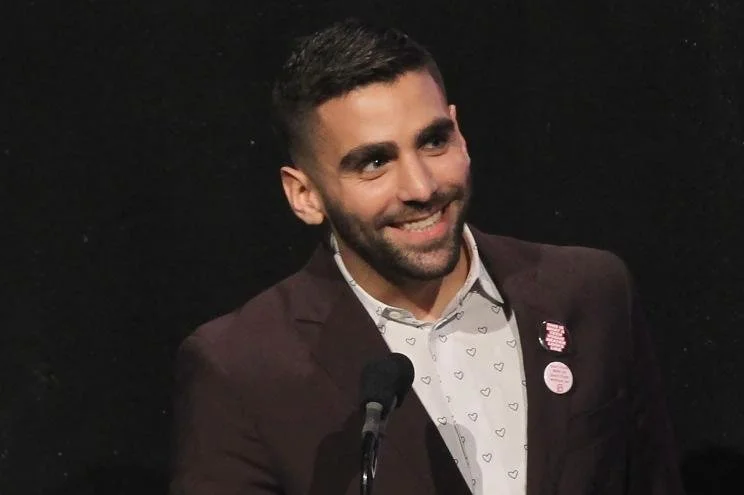A man with dark hair and beard, wearing a white shirt with small hearts, a dark blazer with pins, speaking into a microphone against a black background.