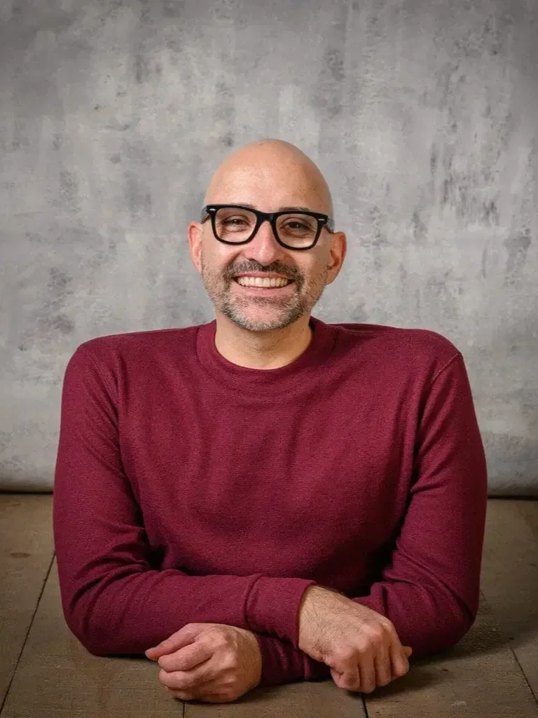 A smiling man with a beard and glasses, wearing a maroon long-sleeve shirt, sitting with his arms crossed on a wooden table in front of a gray wall.