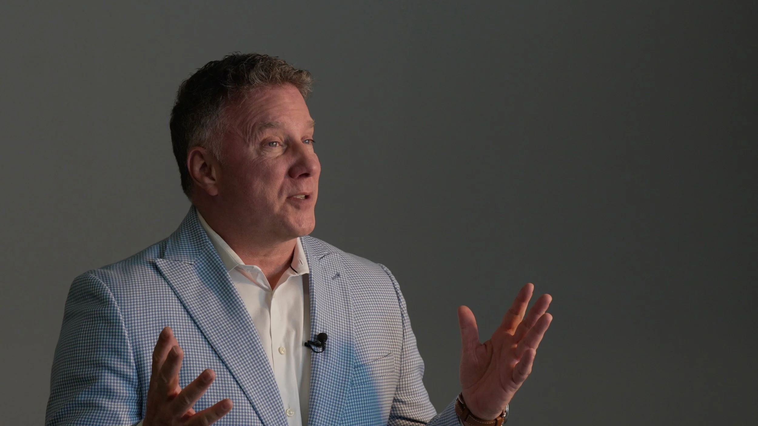 A middle-aged man with light brown hair, wearing a light-colored checked blazer and white shirt, is speaking and gesturing with his hands in front of a plain gray background.