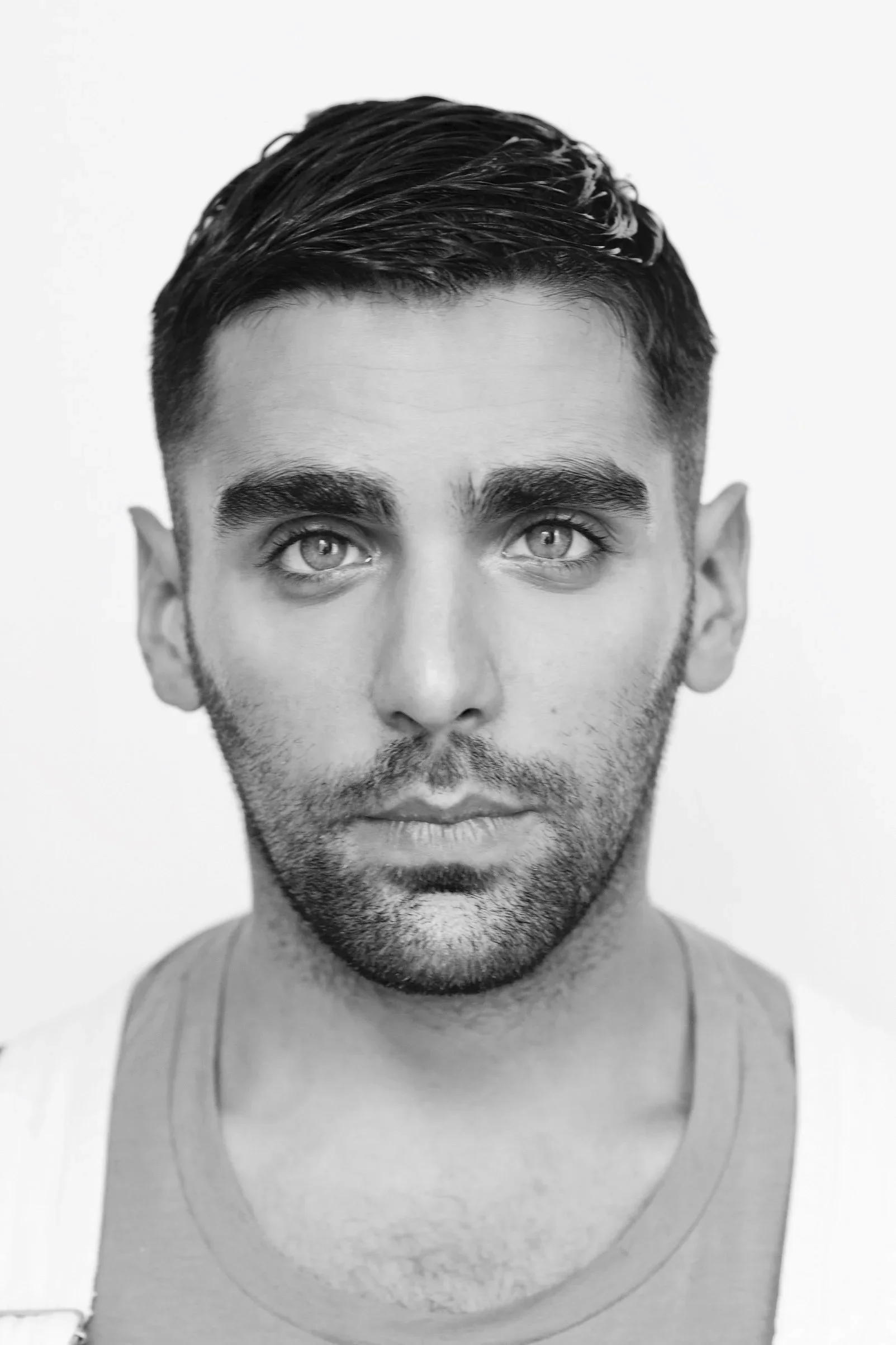 Black and white portrait of a young man with styled hair, light eyes, facial hair, and wearing a tank top.