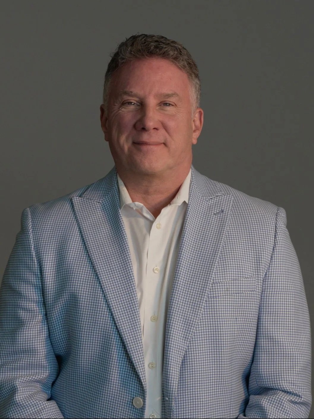 A middle-aged man with short, wavy hair wearing a white dress shirt and a light-colored checkered blazer, standing against a plain gray background.