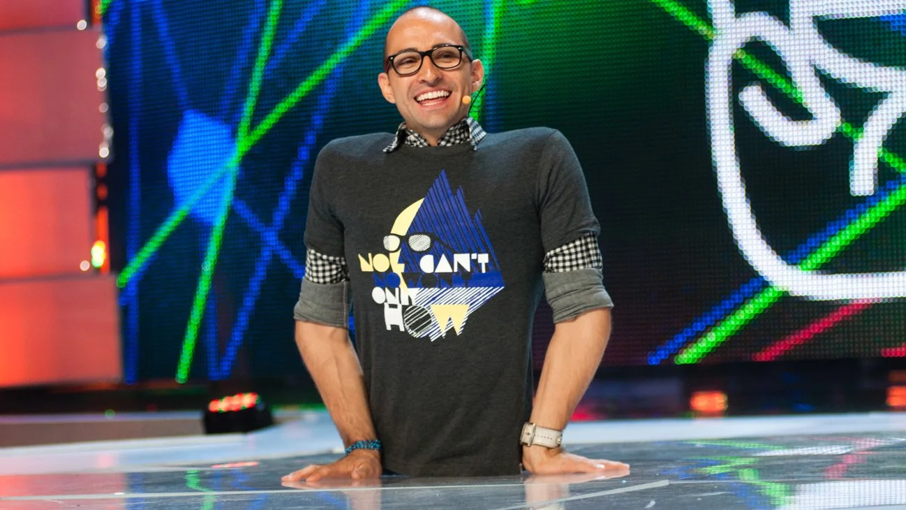 A man with glasses, a long-sleeve gray shirt with rolled-up sleeves, and a graphic T-shirt underneath, smiling and leaning on a reflective surface with a digital stage background behind him.