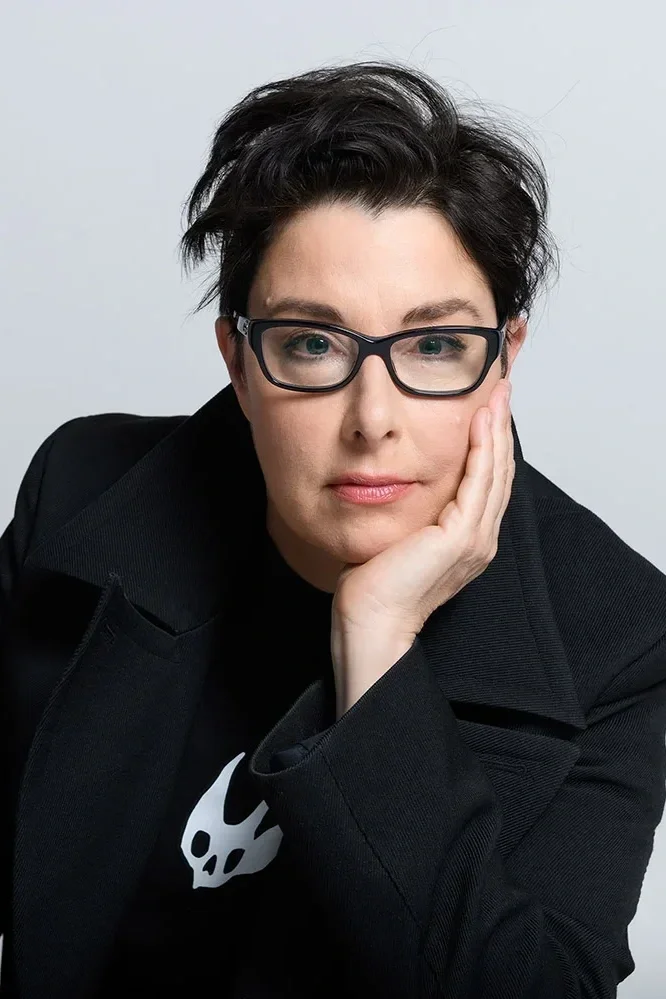 Portrait of a person with short black hair, wearing glasses, a black blazer, and a black T-shirt with a white skull logo, resting their face on their hand, against a light gray background.