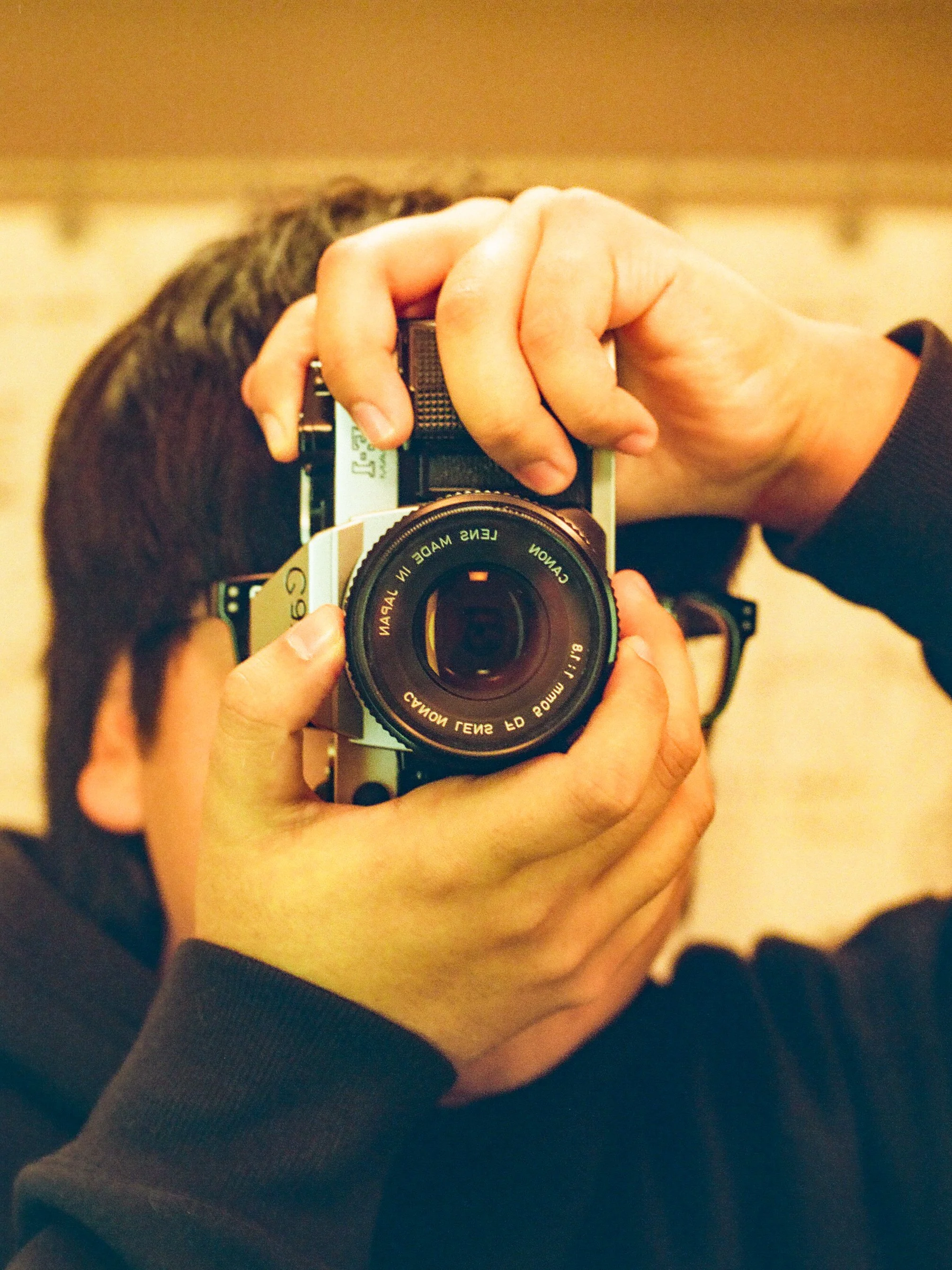 A person taking a selfie with a vintage film camera, focusing on their hand holding the camera and their head, with their dark hair visible.