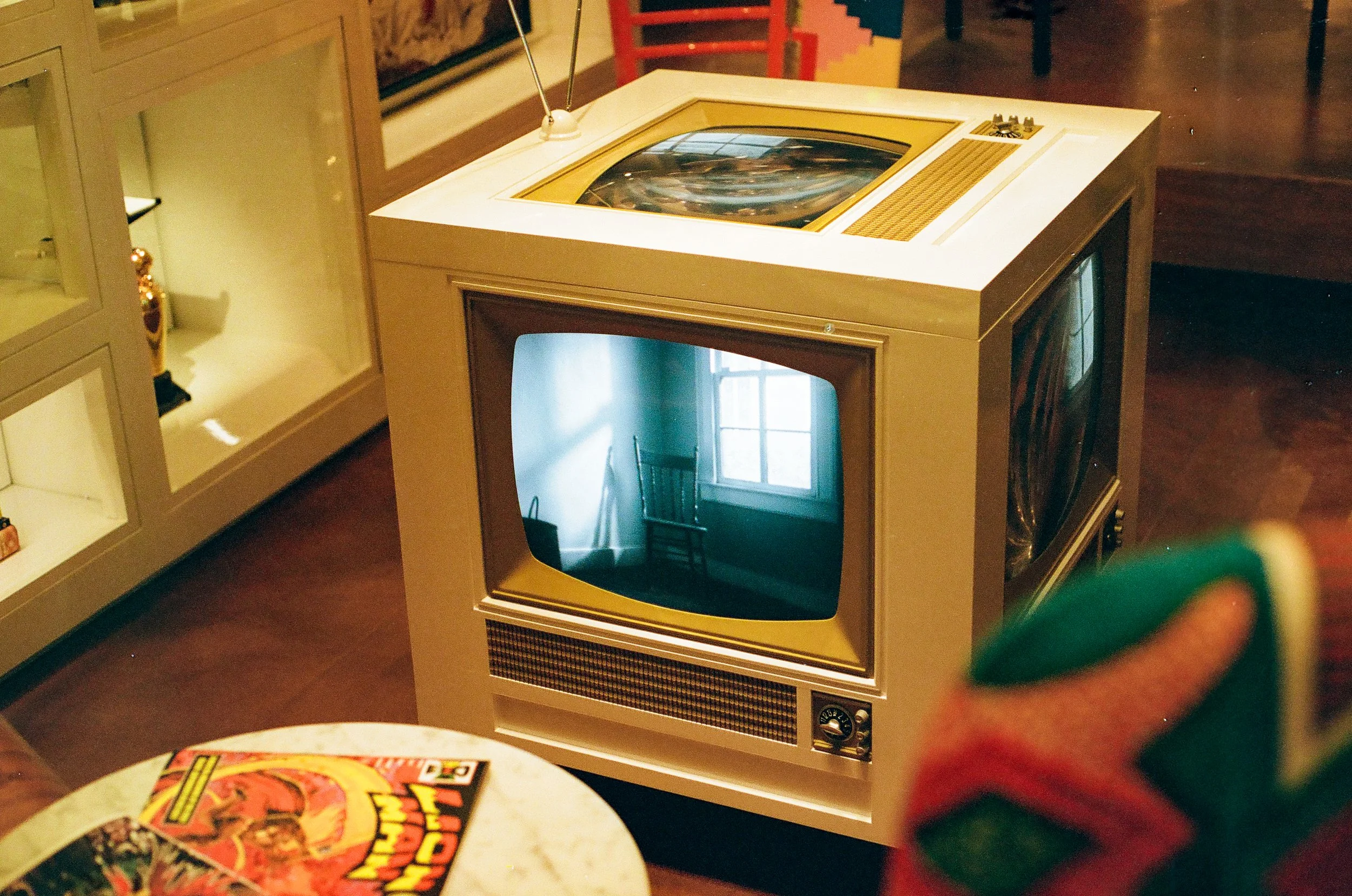 A vintage television set with a rounded screen and dials, placed on a table. In the background, there are pictures and a window. In the foreground, part of a comic book or magazine is visible.