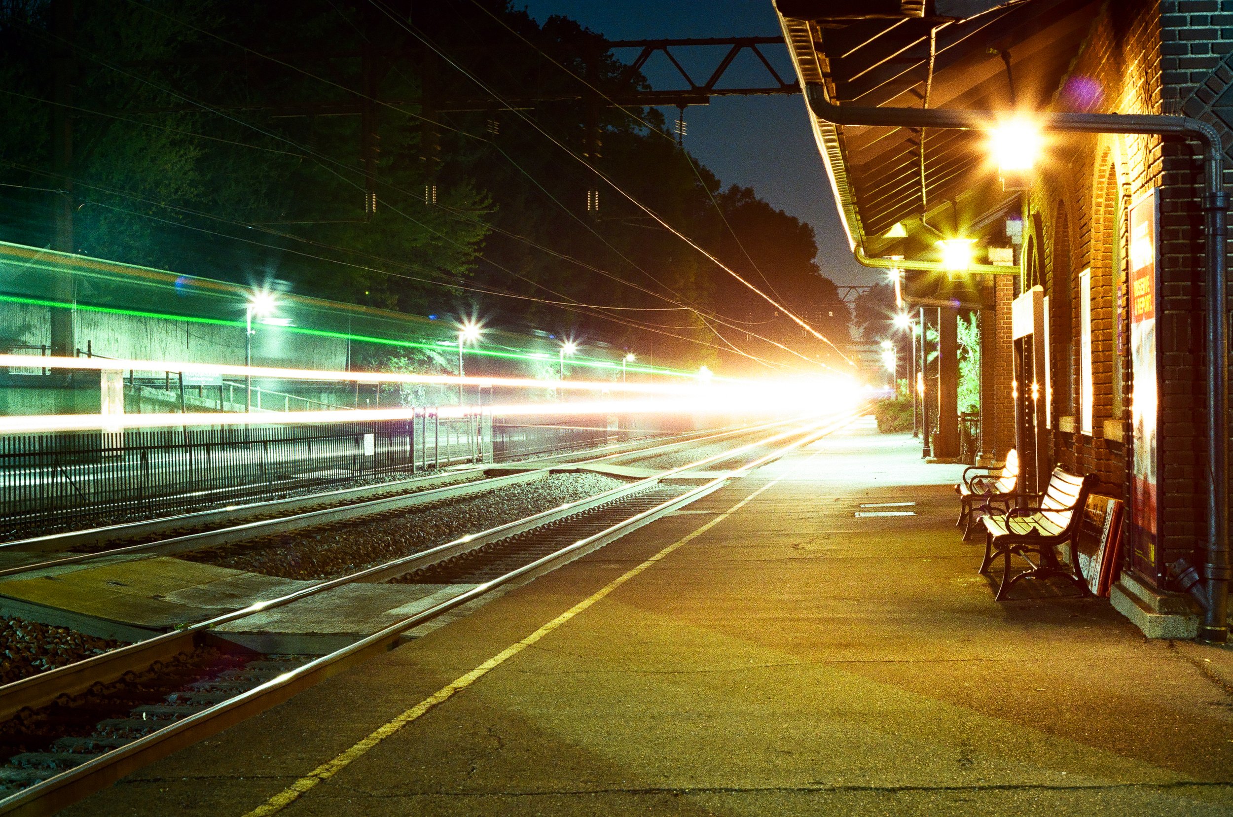 Night scene at a train station with glowing lights, train tracks, and benches along the platform.