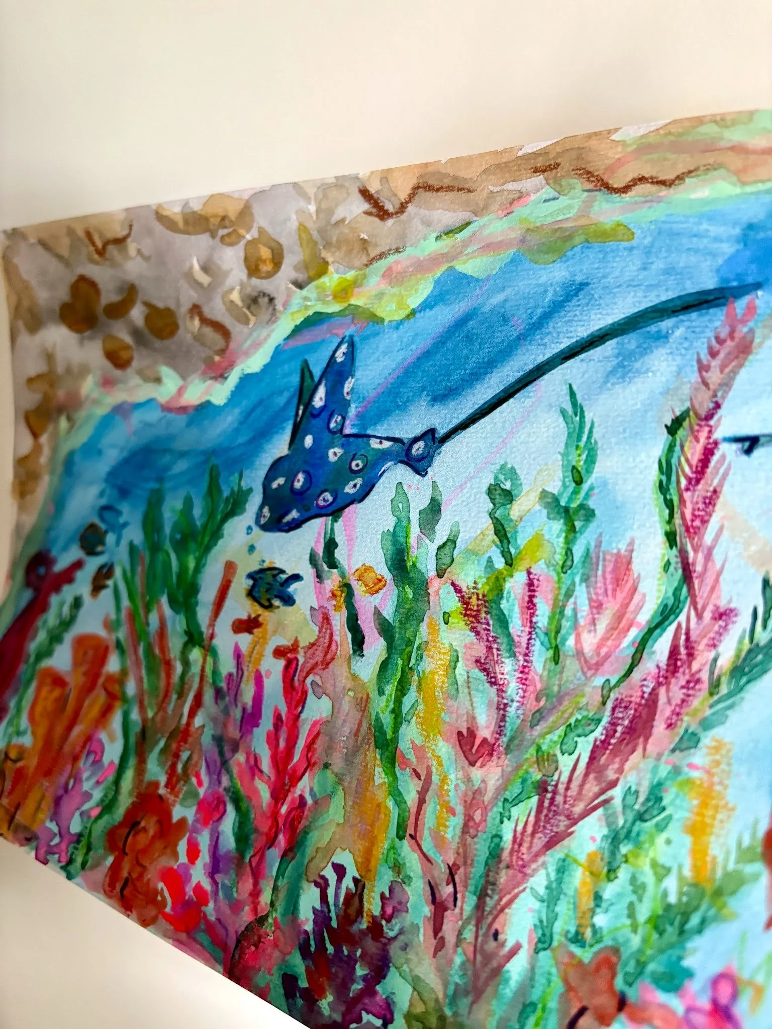 Colorful watercolor painting on paper featuring a blue unicorn with a star on its head, surrounded by green grass and pink plants, with a brown and green landscape in the background.