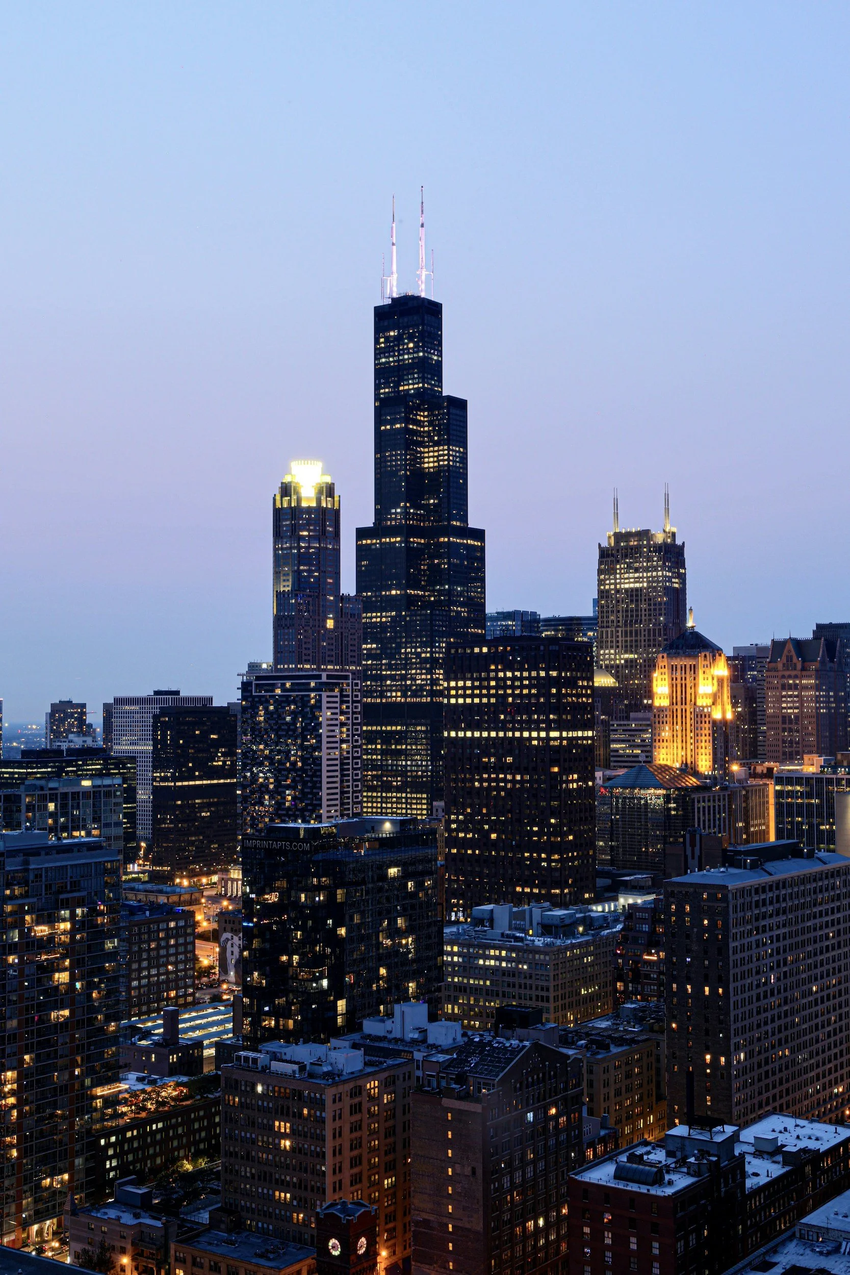A city skyline at dusk with the Willis Tower (Sears Tower) in Chicago, illuminated buildings, and a light blue sky.