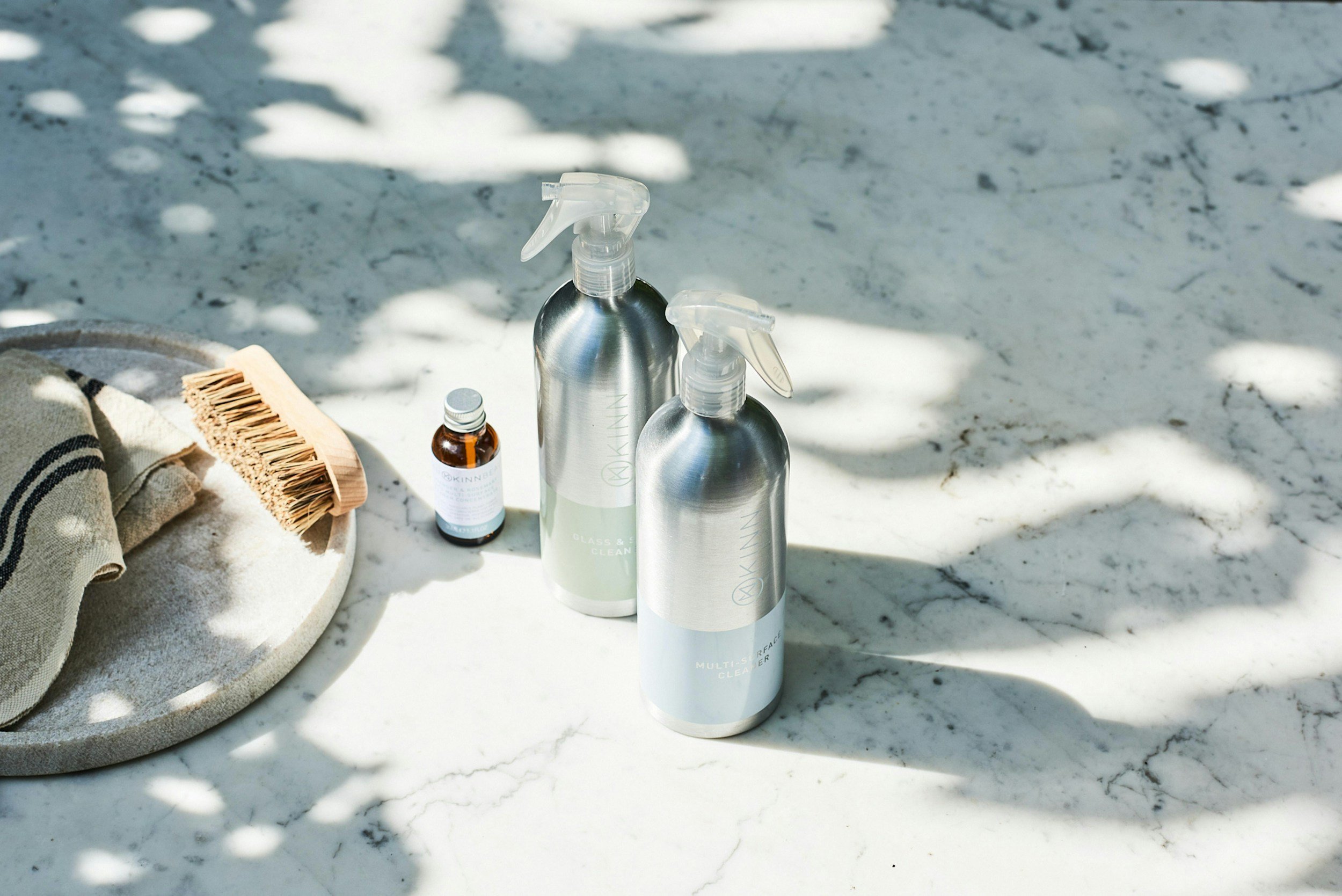Cleaning spray bottles, a small amber bottle, a scrubbing brush, and a towel on a white marble surface outdoors with dappled sunlight and shadows.