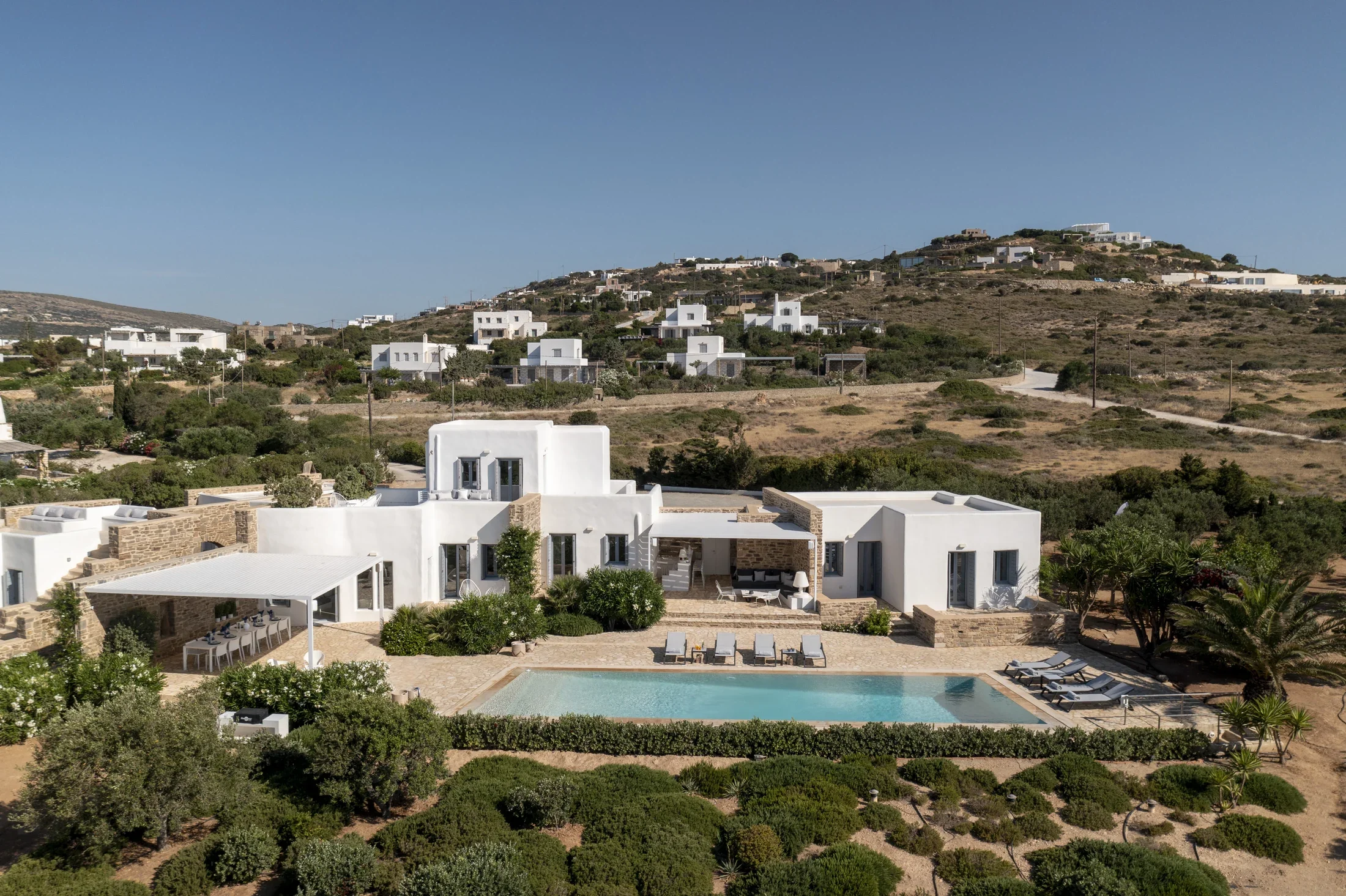 A hillside with white modern houses, some with flat roofs, overlooking a pool area with lounge chairs and greenery in a Mediterranean setting under a clear blue sky.