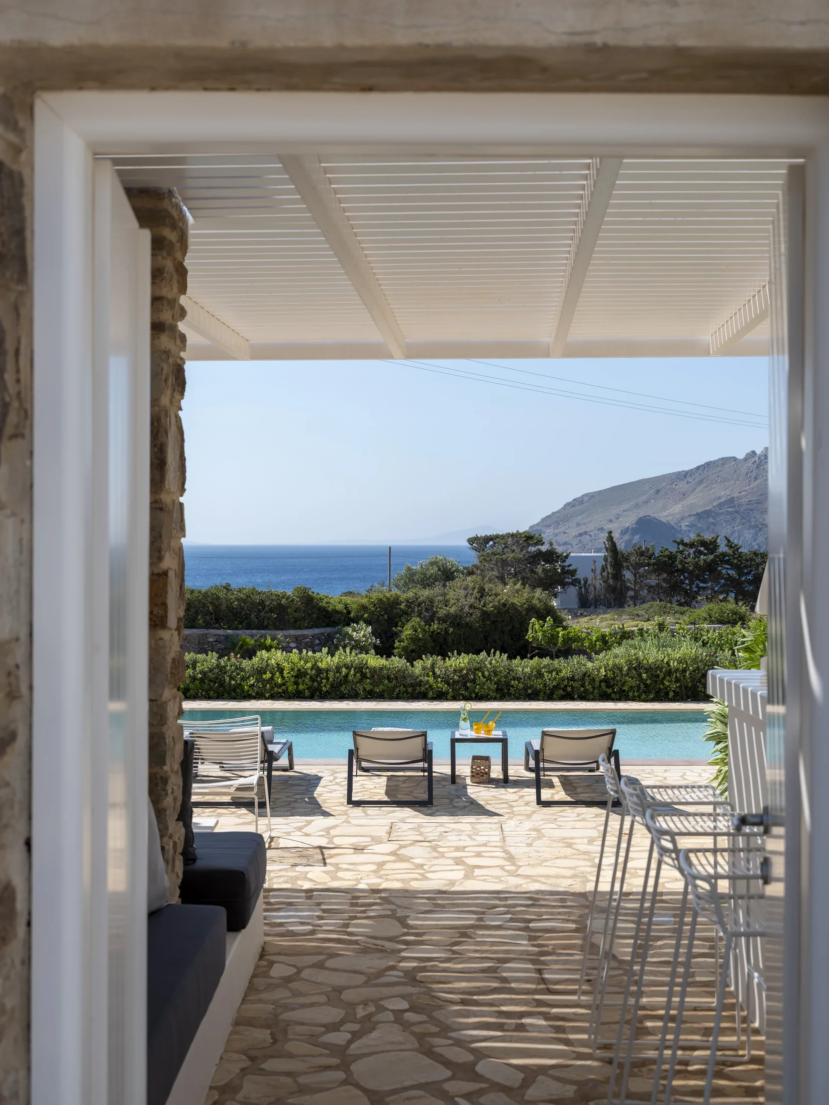 View through a doorway of a poolside patio with lounge chairs, a small table with drinks, vienyards, and the background of a beach in Paros island.