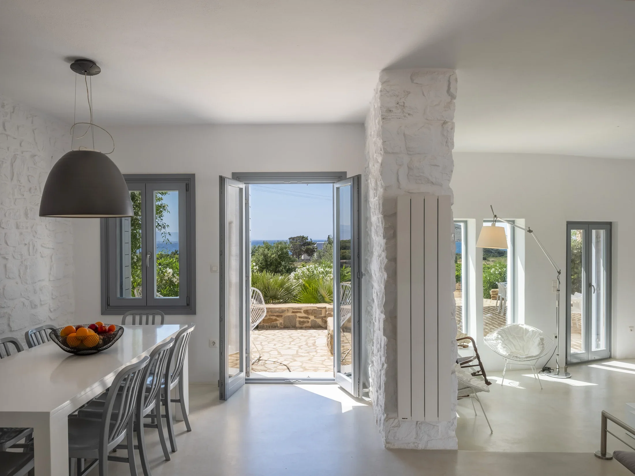 Interior view of a bright, white room with large open French doors leading outside, revealing greenery and a glimpse of the ocean. There is a white dining table with metal chairs, a hanging black pendant light, and a white stone wall dividing the spa